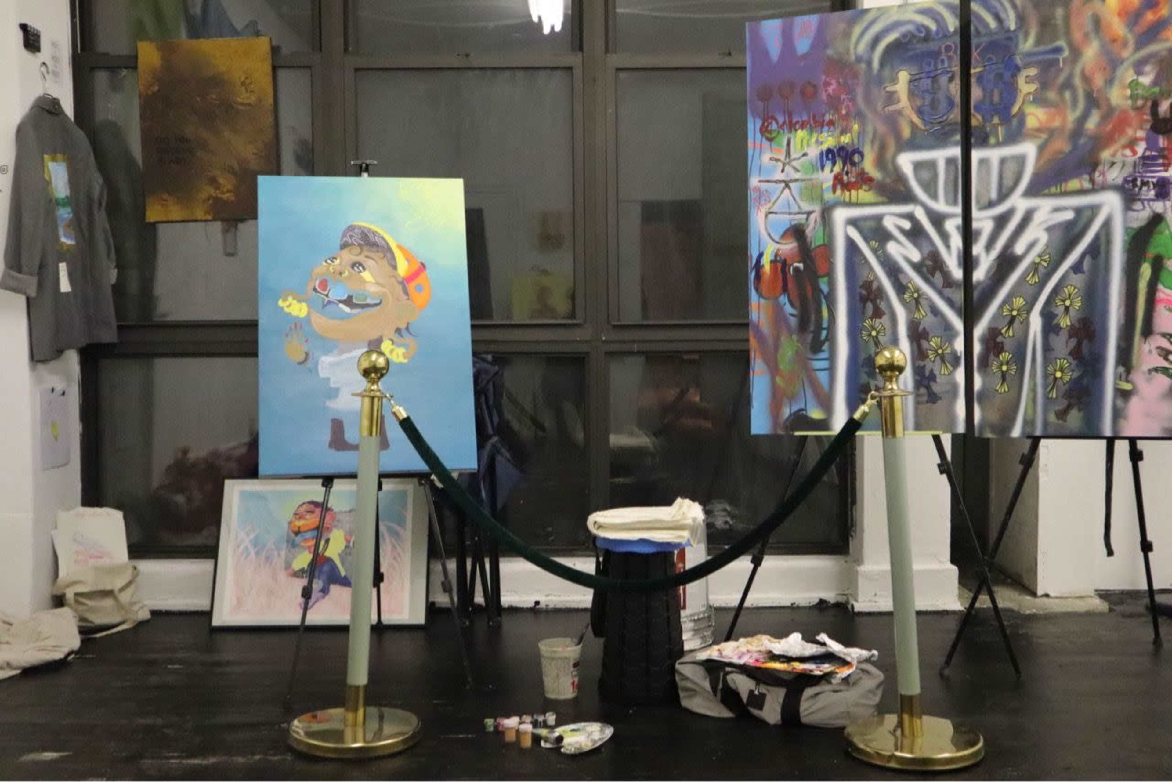 The image shows an art setup in a gallery featuring two paintings on easels, a bucket of paint, and various art supplies on the floor, with a wall displaying colorful artworks in the background.