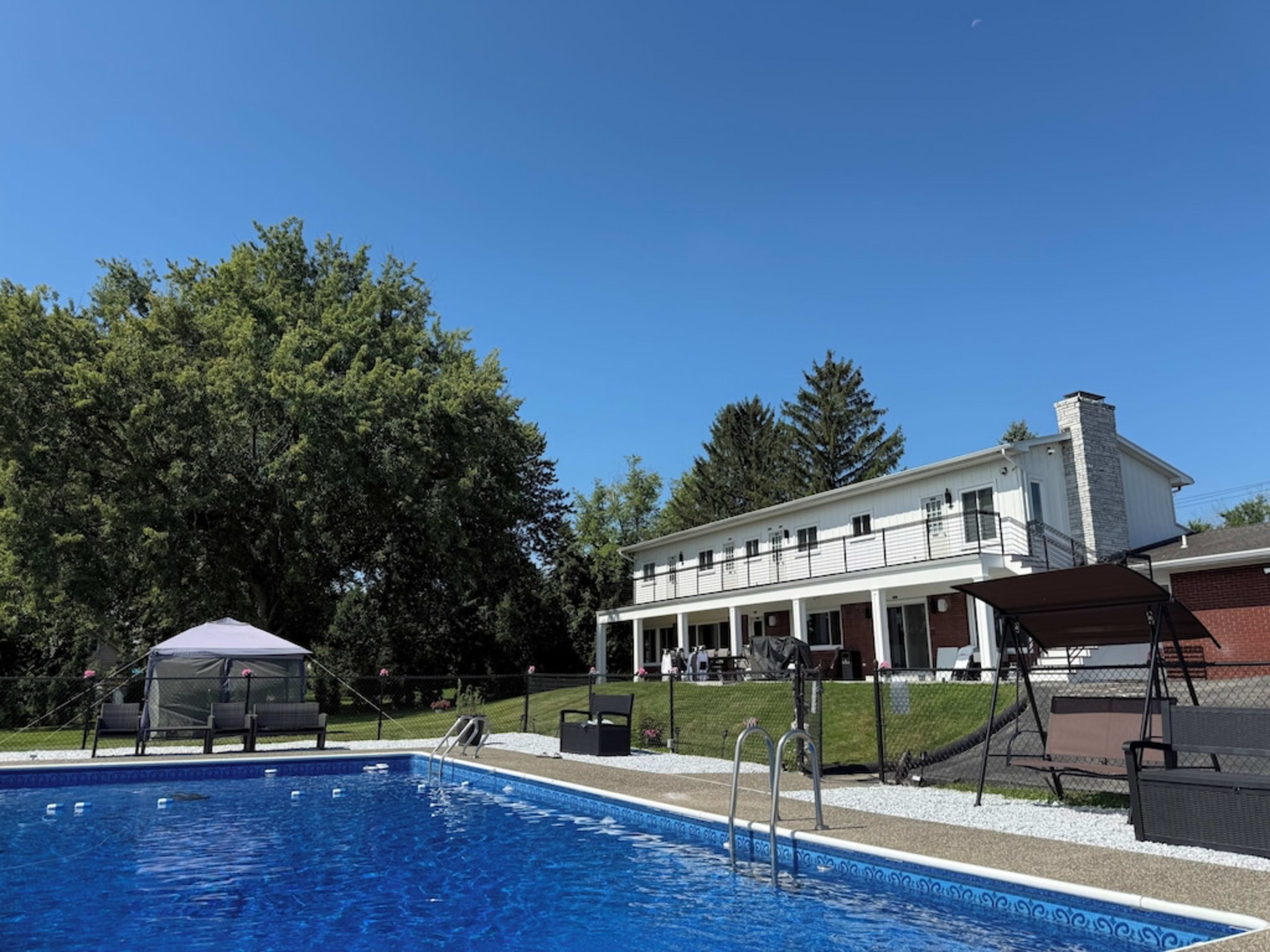 The image shows a clear blue swimming pool in the foreground, with a two-story house and green trees in the background under a sunny sky.