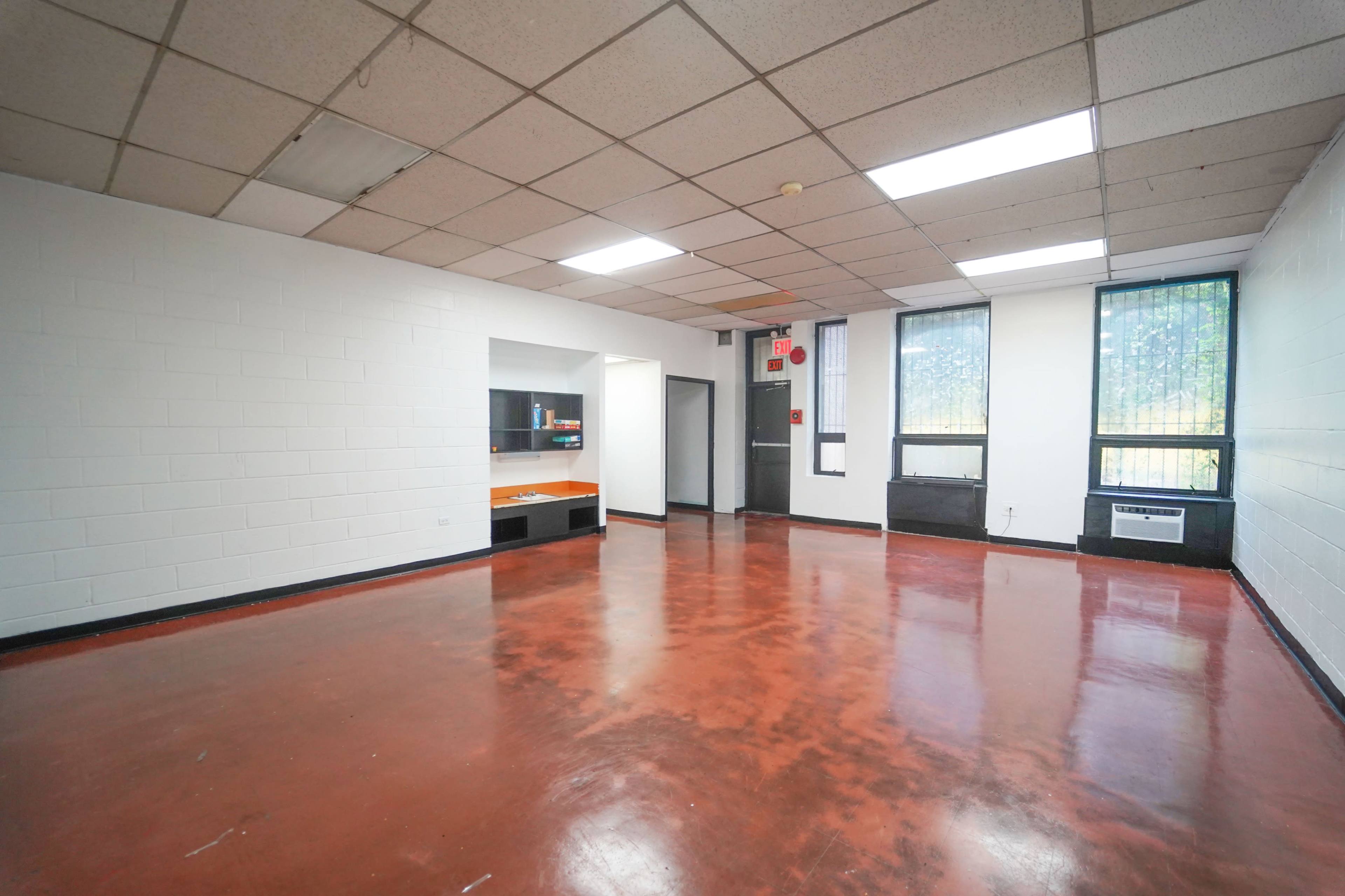 The image shows an empty room with a polished red concrete floor, white walls, and large windows on one side.