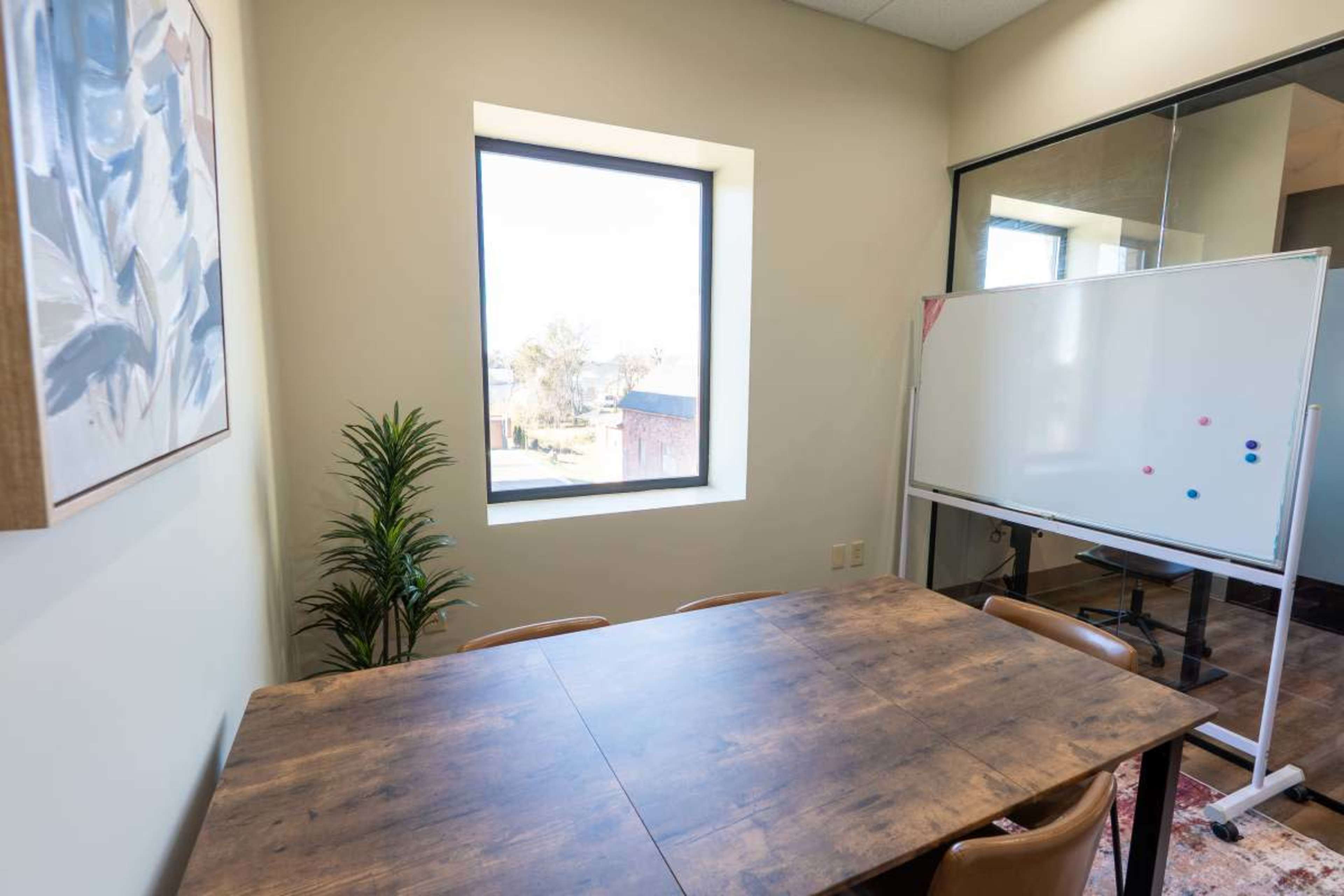 A meeting room features a wooden table, four chairs, a whiteboard, and a large window with a view of the outside.