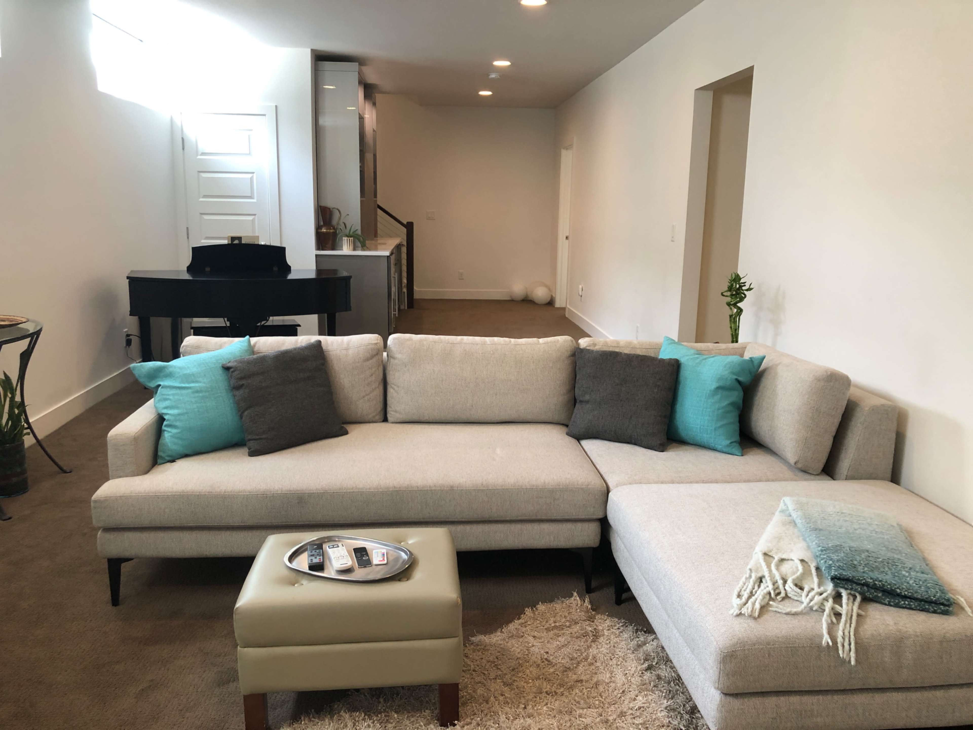 The image shows a modern living room with a light-colored sectional sofa, a small coffee table, and a grand piano in the background.