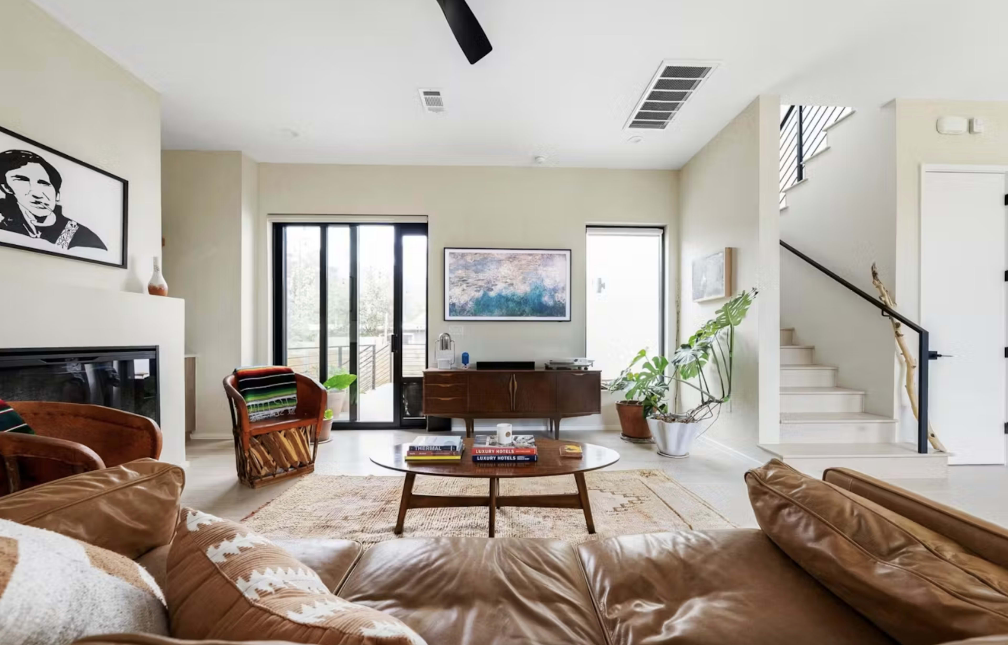 The image shows a modern living room featuring a brown leather sofa, a dark wooden coffee table, a television on the wall, and stairs leading to an upper level.
