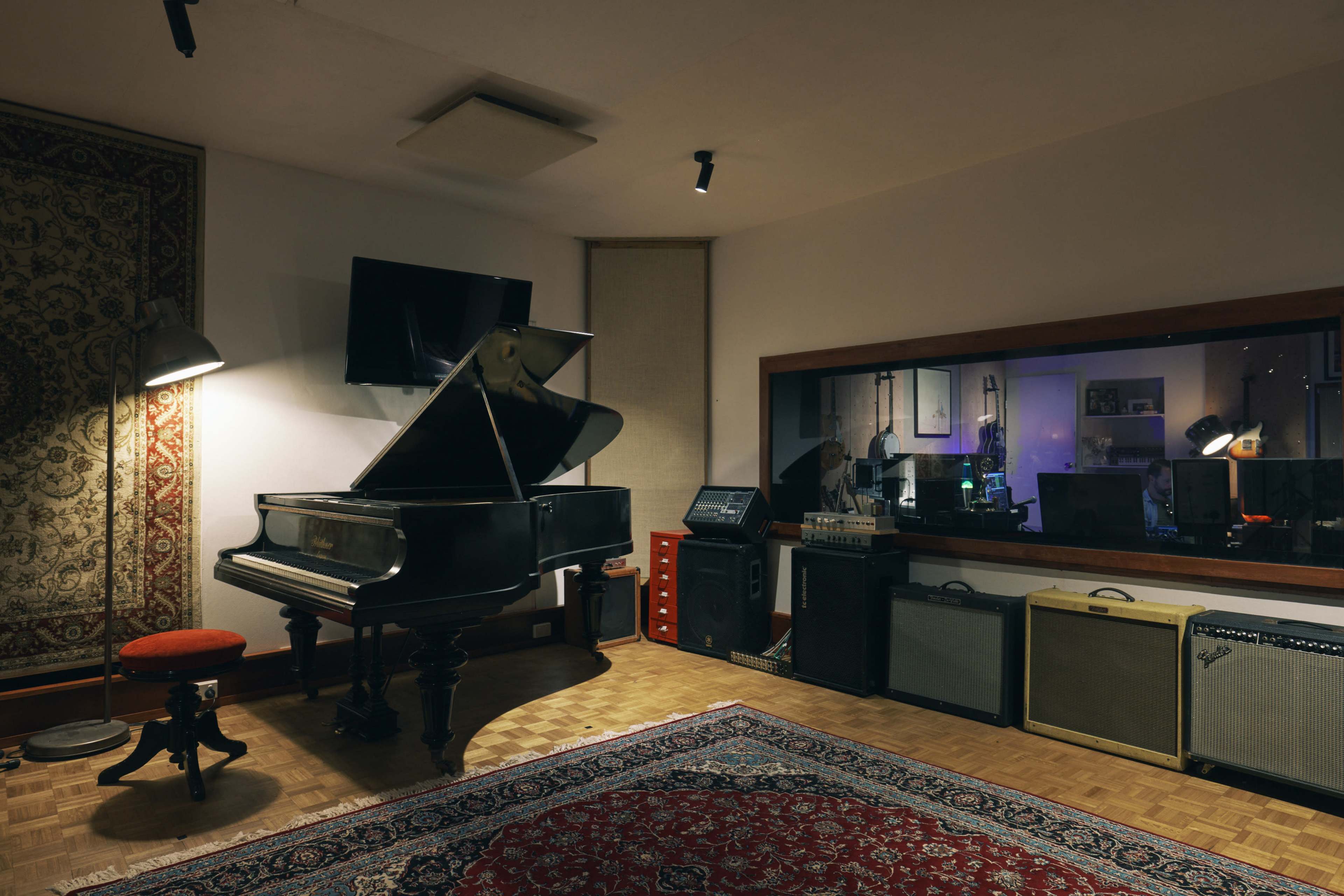 The image shows a music studio with a grand piano, recording equipment, and a plush rug on a wooden floor.