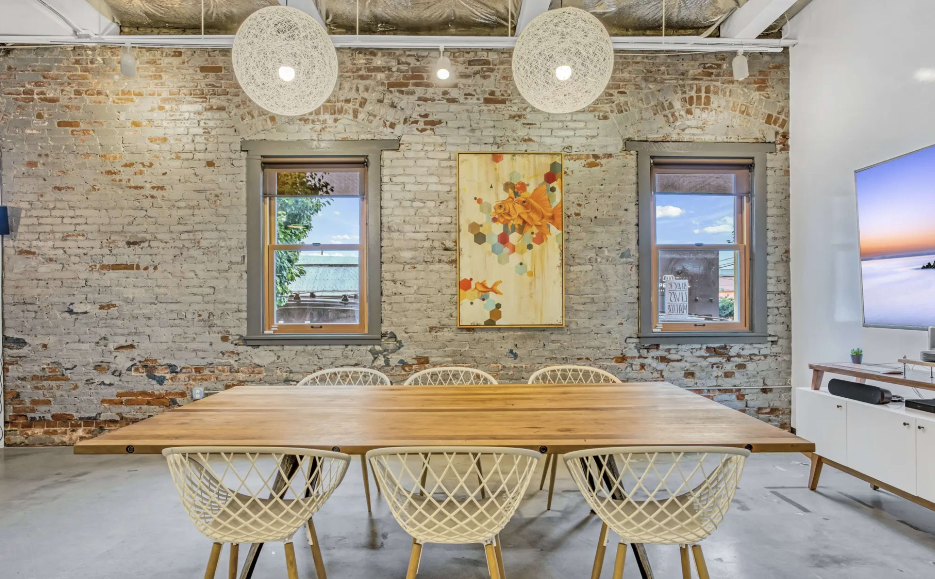 The image shows a dining area with a wooden table surrounded by white wicker chairs, set against a backdrop of exposed brick walls and two windows.