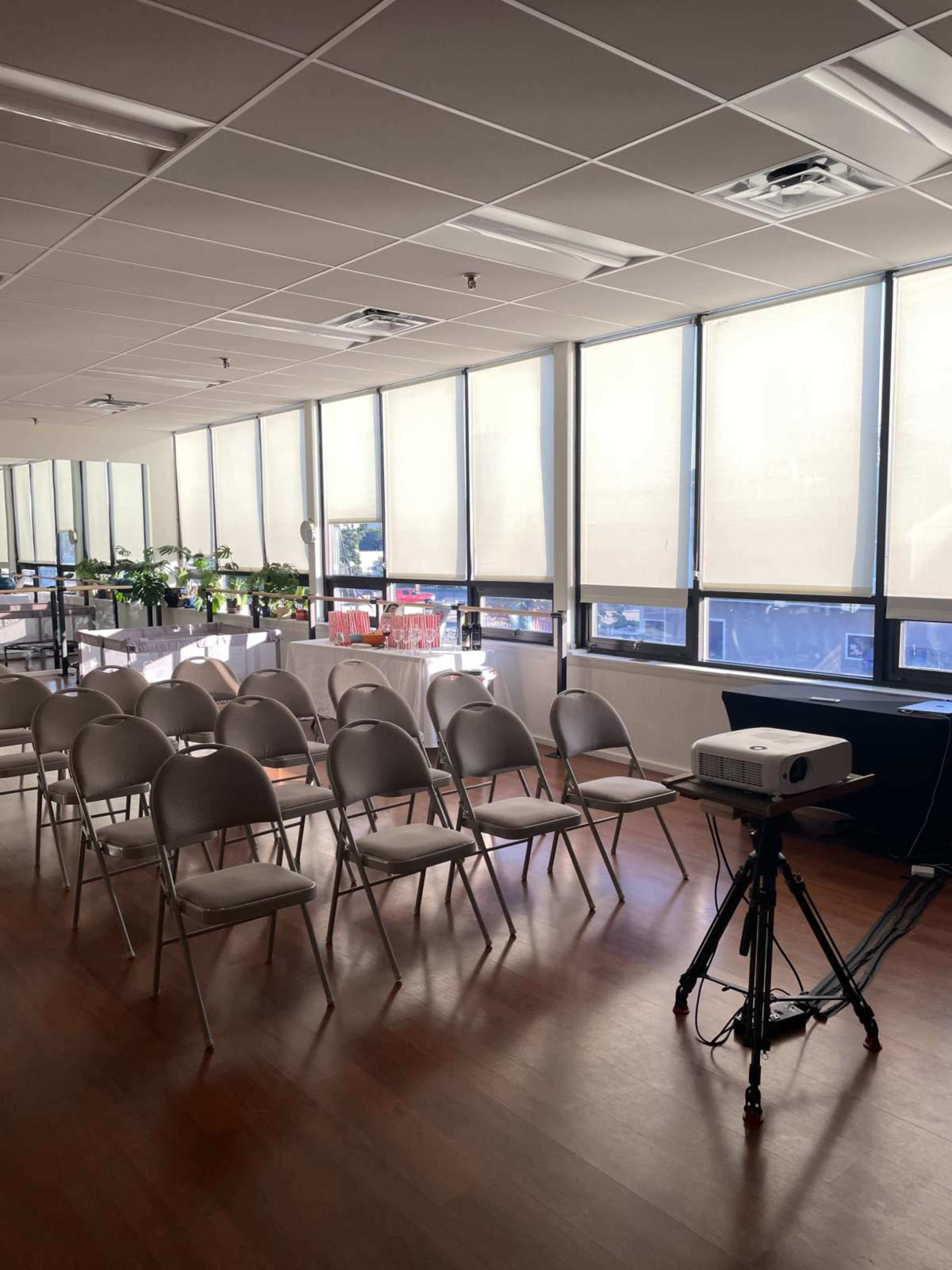 The image shows a room set up for a presentation with rows of empty chairs and a projector on a stand.