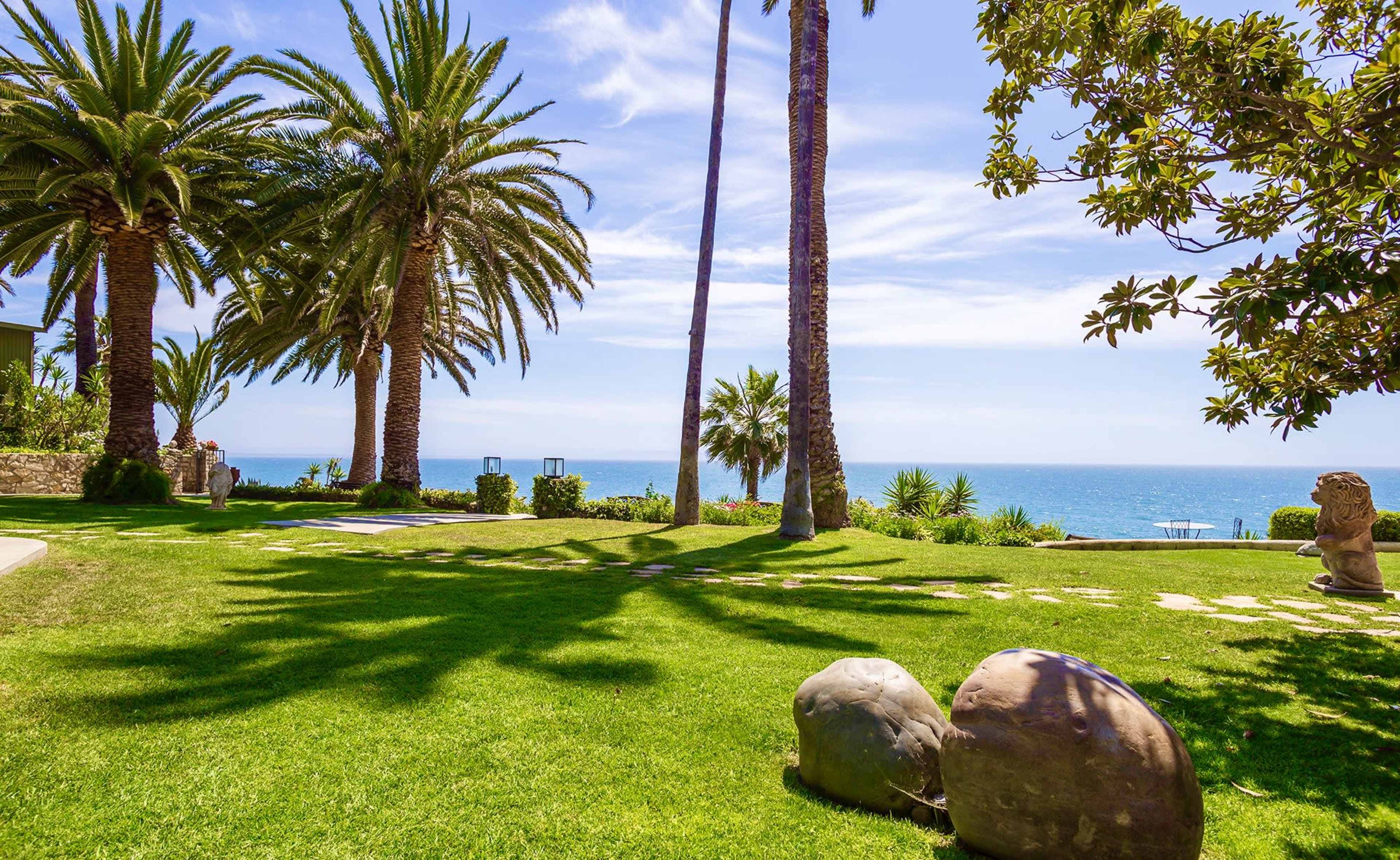 The scene shows a lush green lawn with palm trees and large rocks, overlooking a clear blue ocean under a partly cloudy sky.