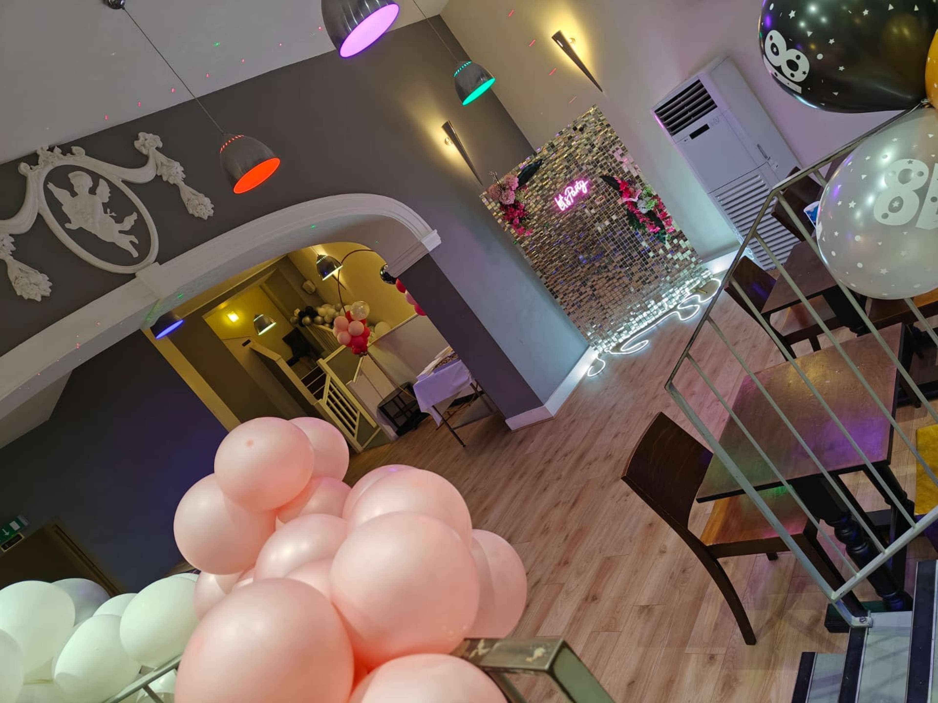 The image shows a decorated indoor venue featuring colorful balloons, a wall with floral arrangements, and a lit-up sign that reads "HAPPY."