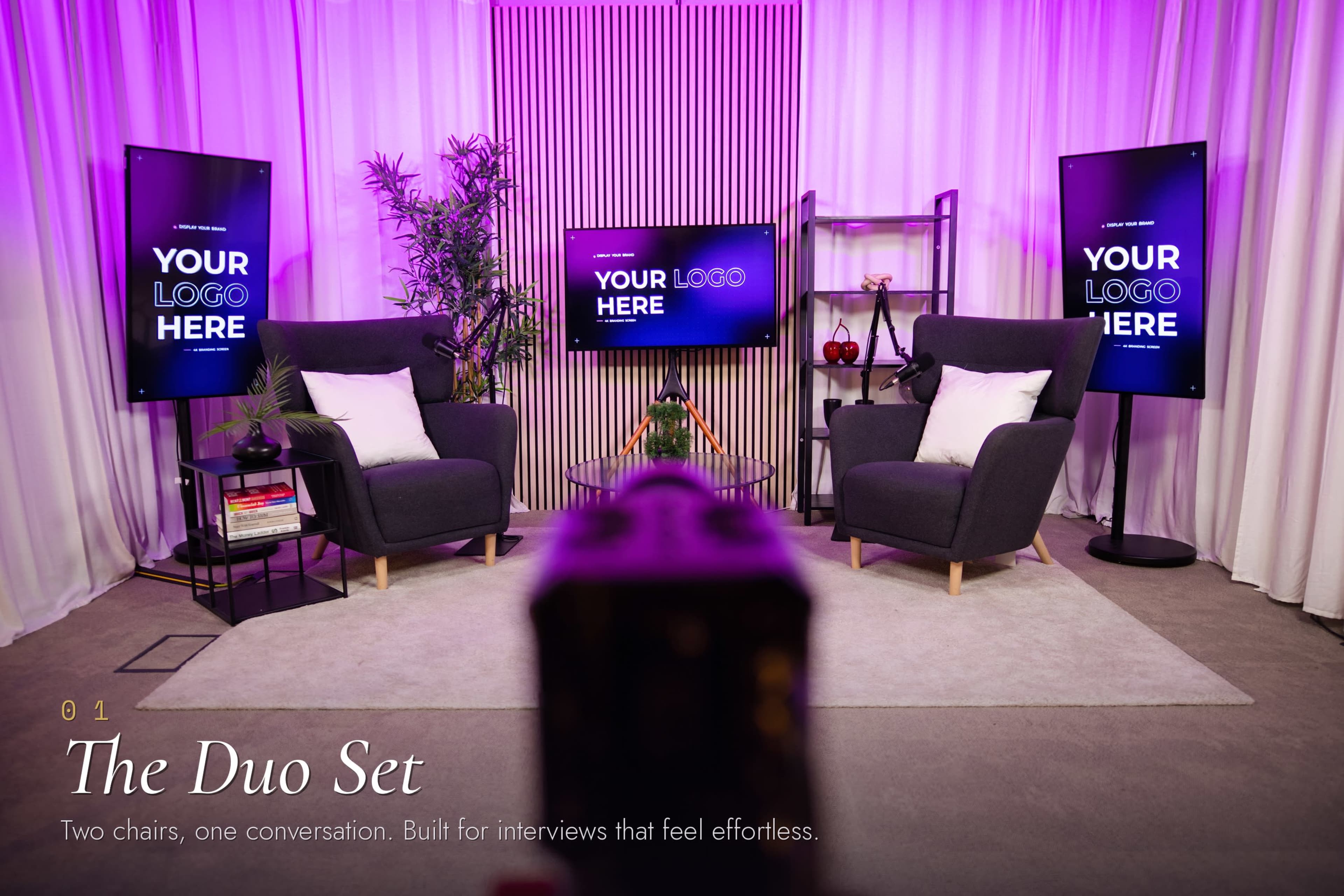 The image features a modern interview setup with two armchairs, a small table, and digital screens displaying customizable logos, all against a backdrop of soft lighting and decorative plants.