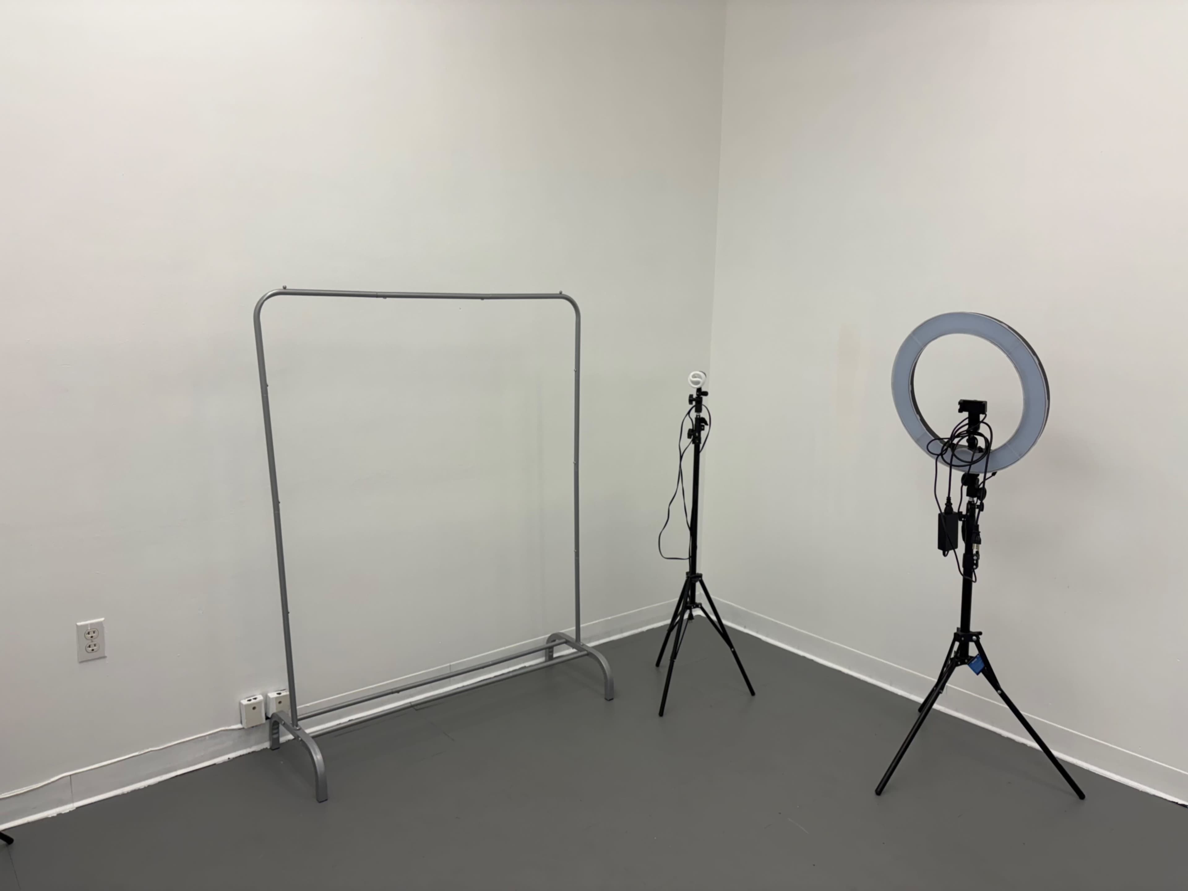 The image shows a plain room with a clothing rack on the left and two light stands, one with a ring light, on the right.