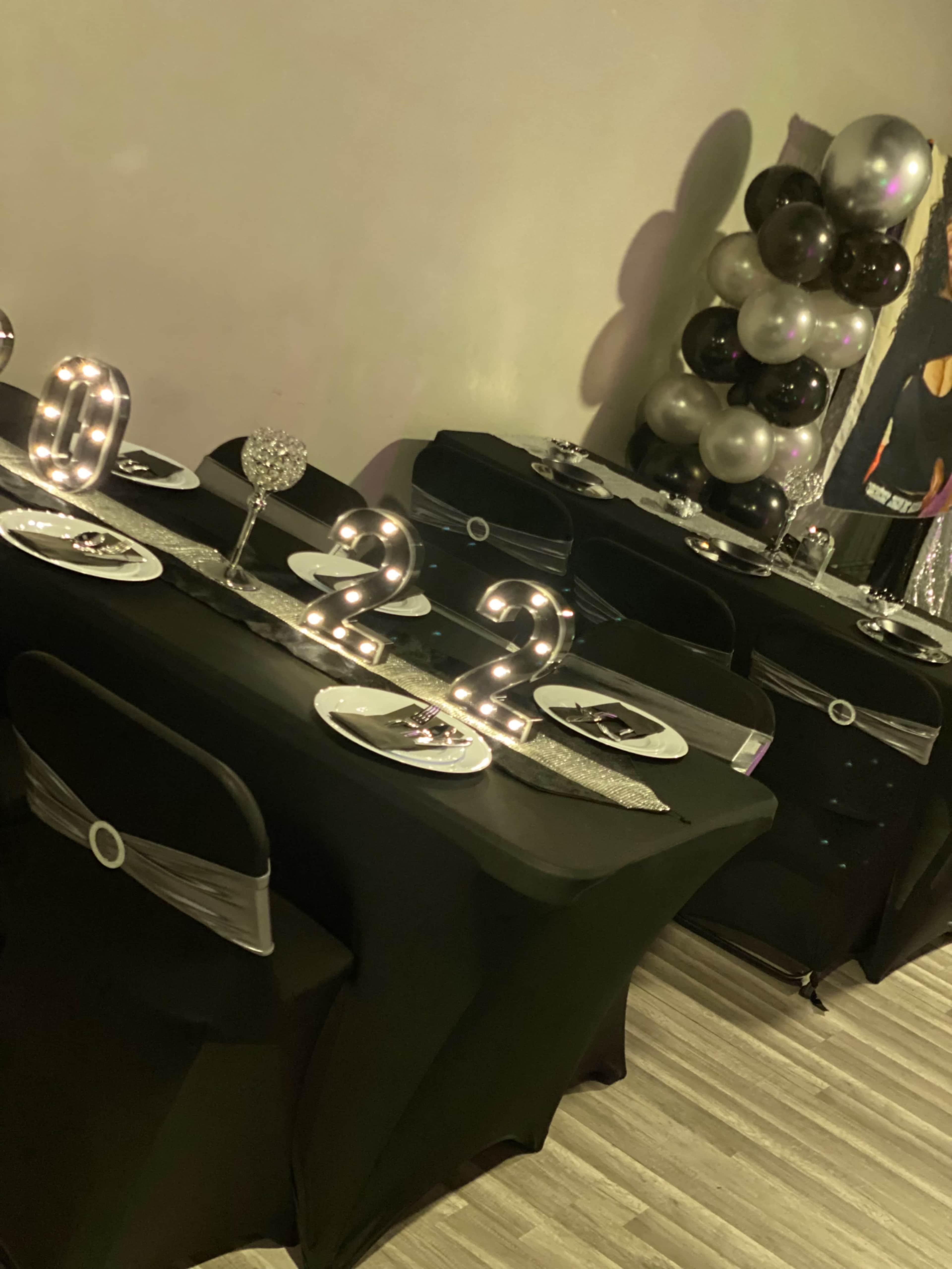 A dining setup features black tables with silver and black decorations, including table numbers illuminated in lights.