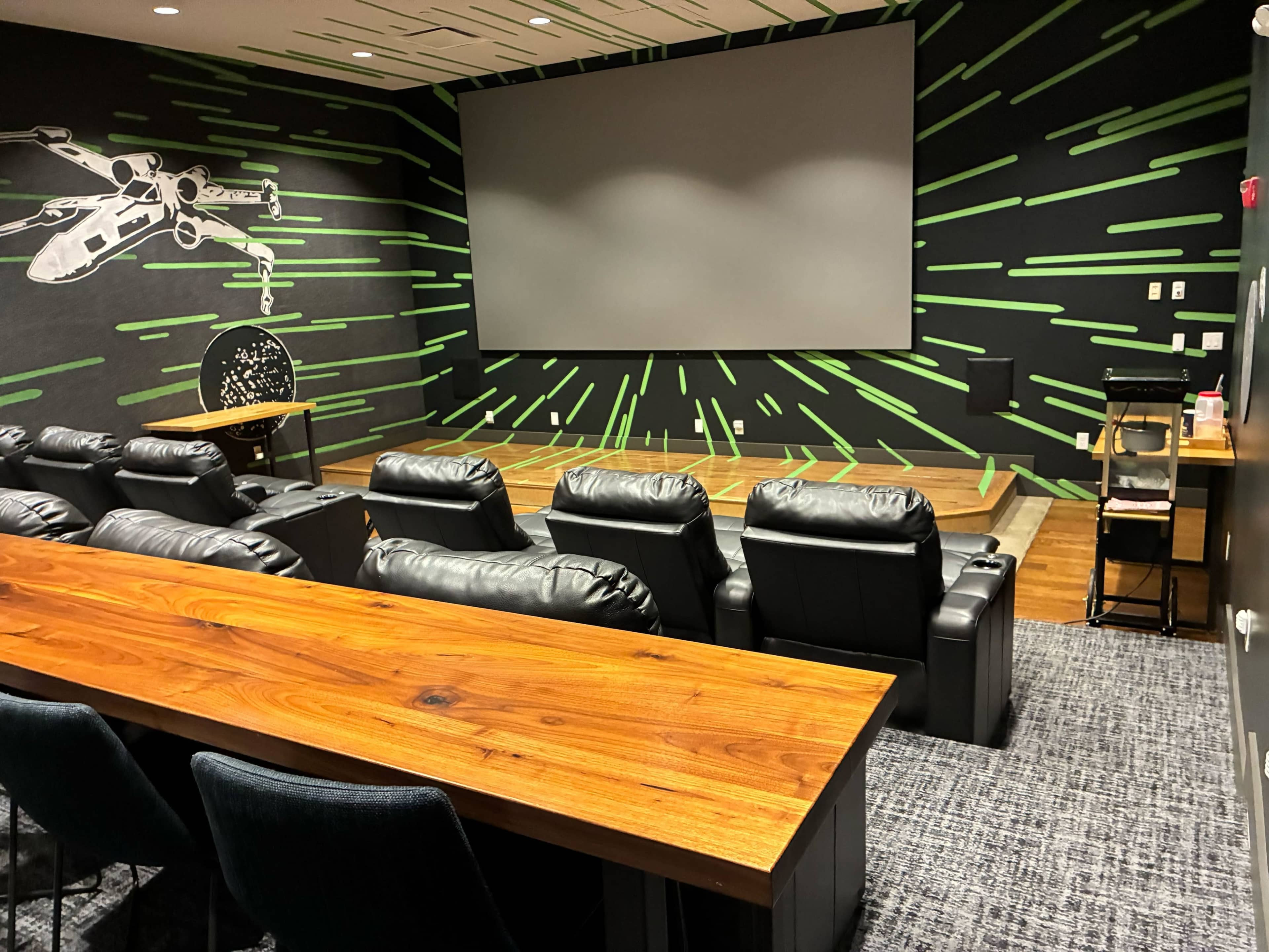 The image shows a home theater setup with black leather reclining seats, a large screen mounted on a wall, and a wooden table in the foreground.