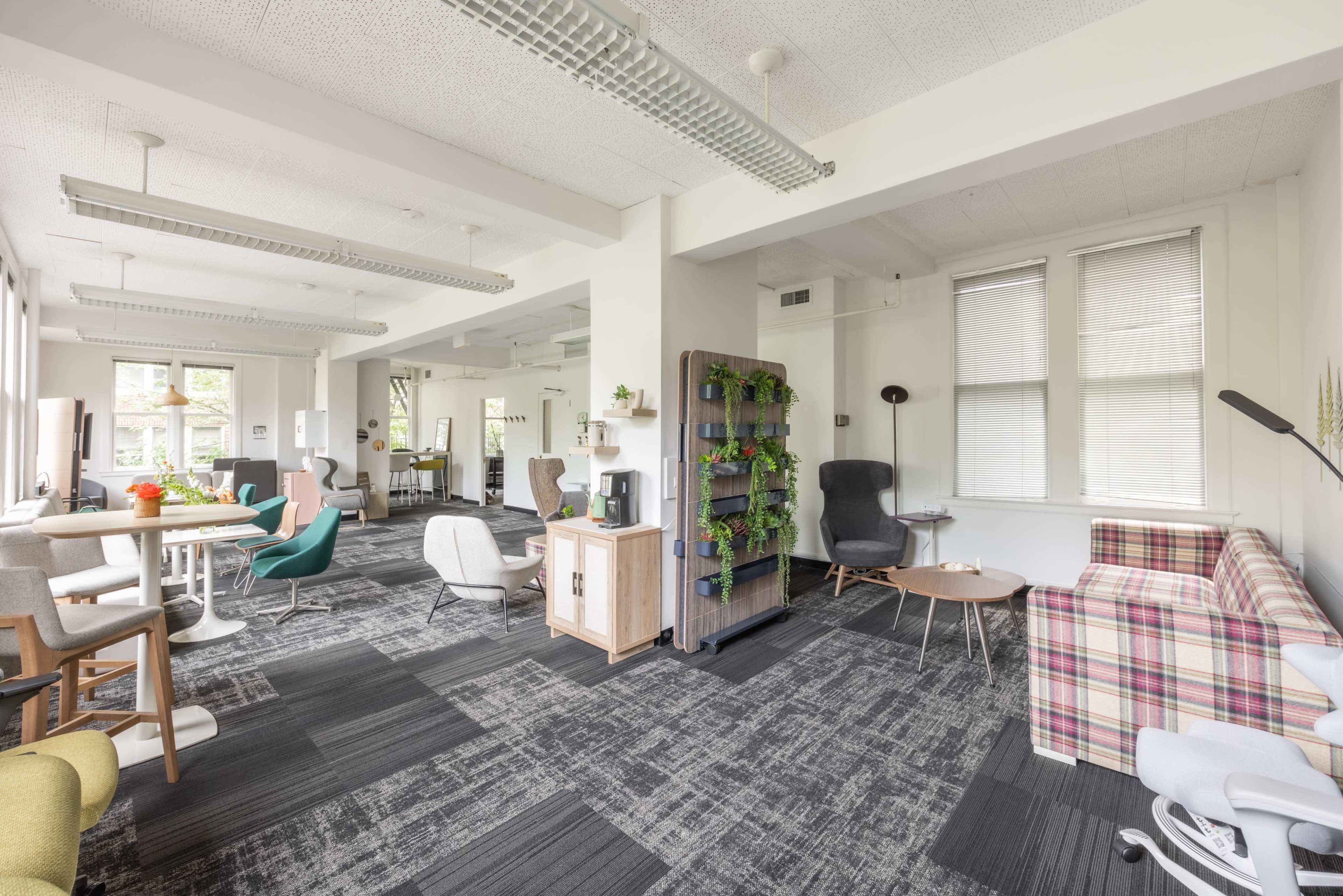 The image shows a spacious, modern open area with various seating options, a bookshelf with plants, and large windows allowing natural light.