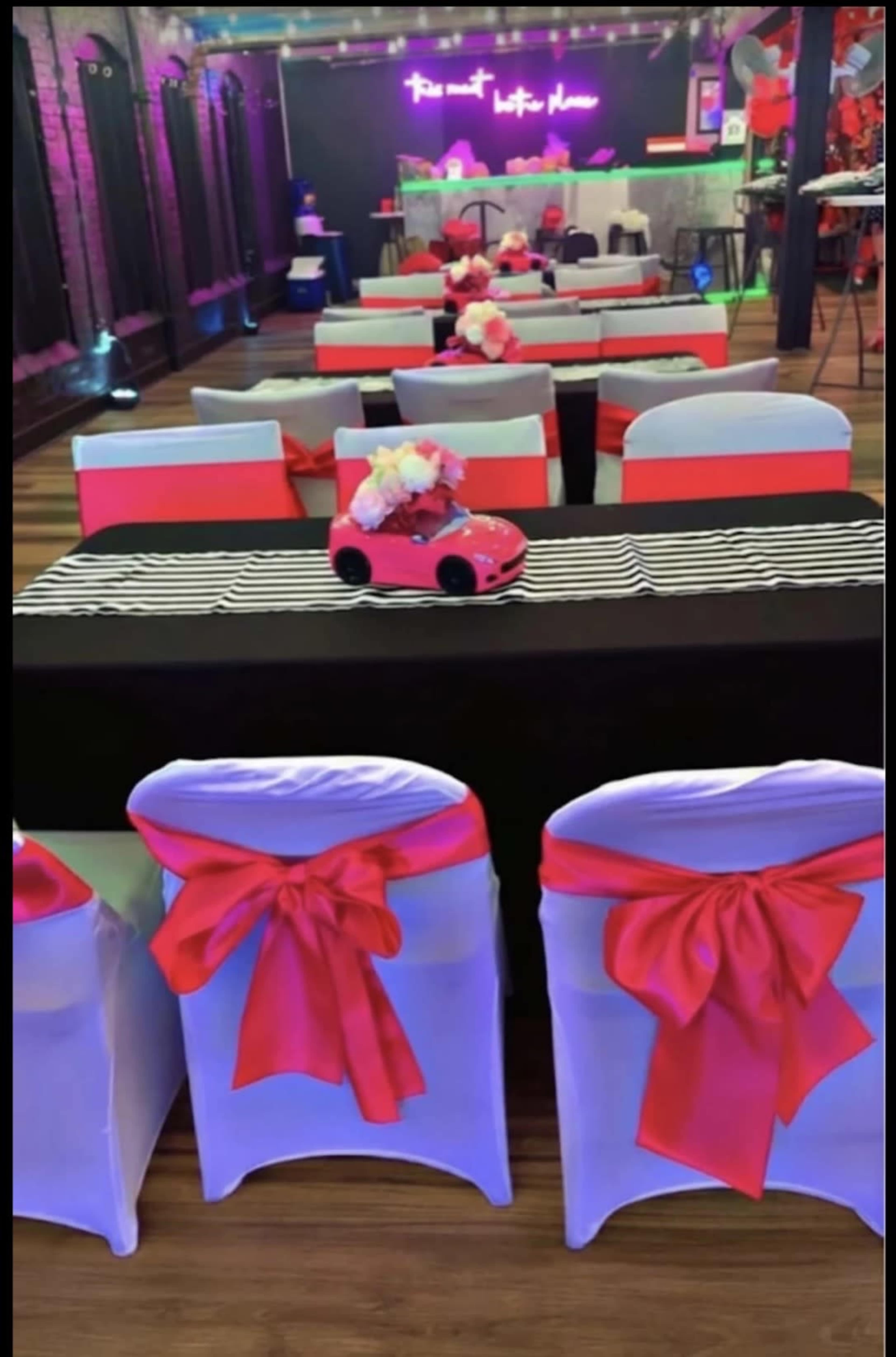 The image shows a decorated event space with tables covered in black and white tablecloths, red sashes on chairs, and a pink toy car as a centerpiece.