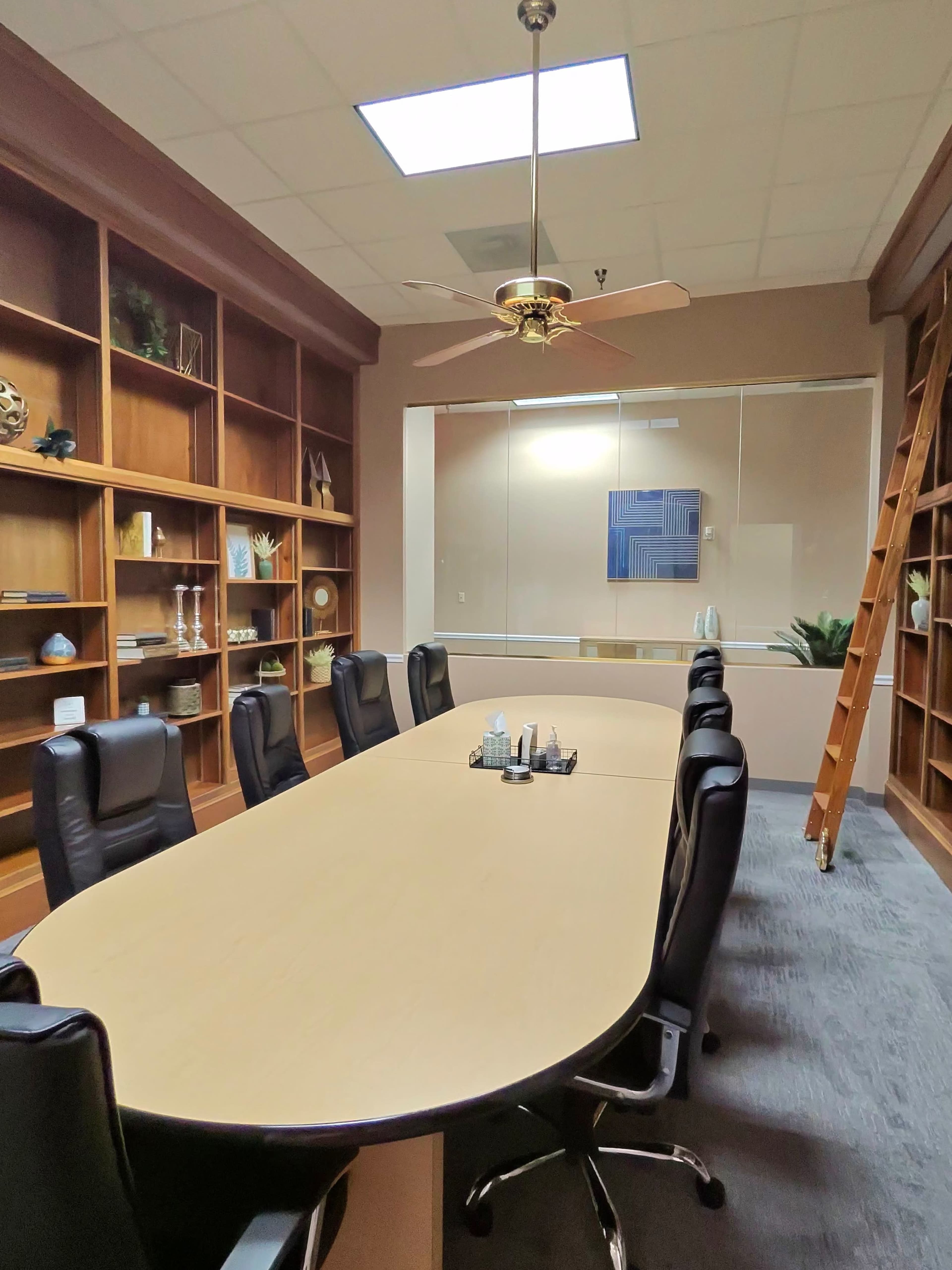 A large conference room features a long oval table surrounded by black chairs, with bookshelves and a glass-walled meeting area in the background.