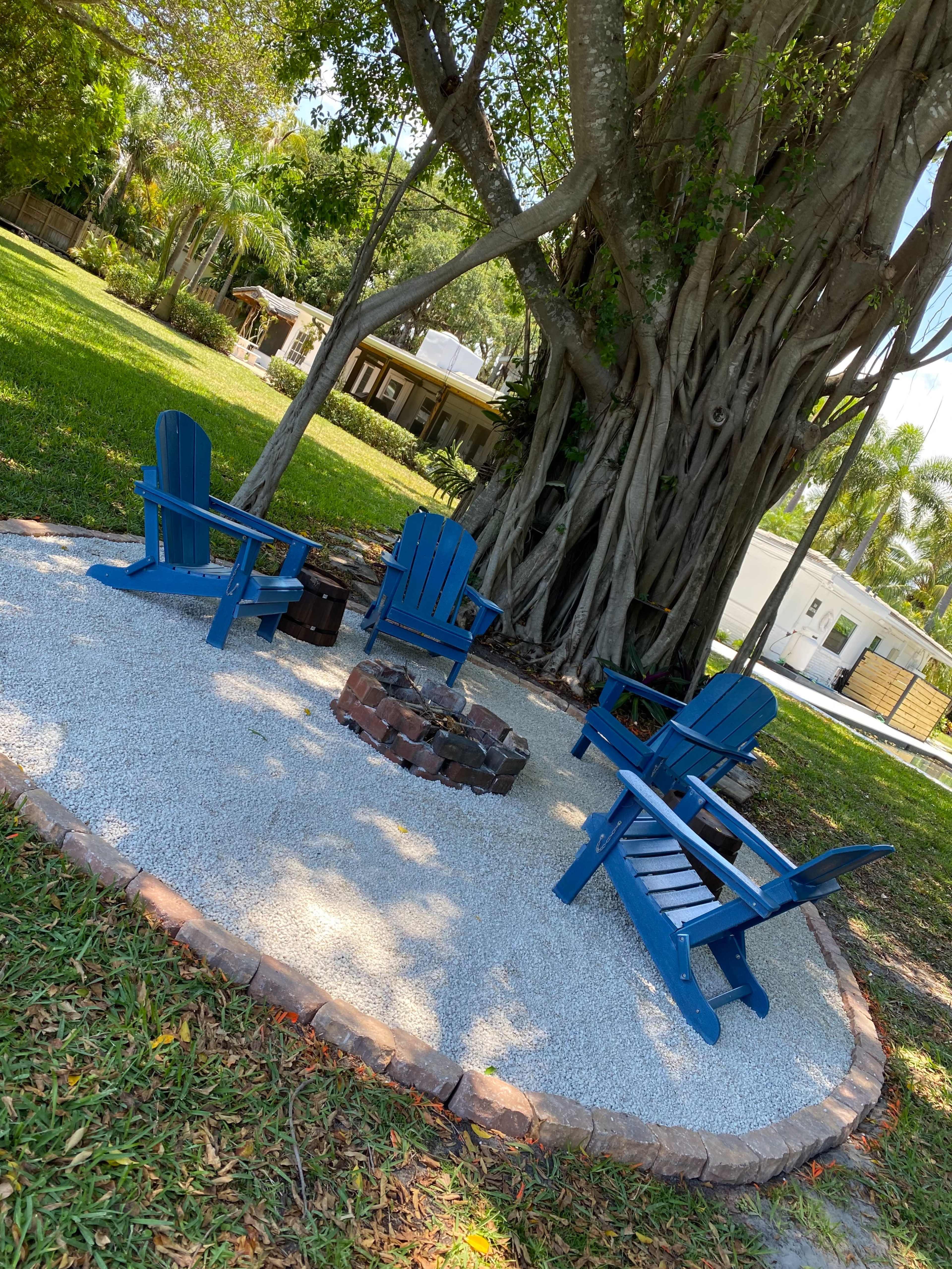 A cozy fire pit area with blue Adirondack chairs is surrounded by gravel and situated under a large tree in a grassy yard.