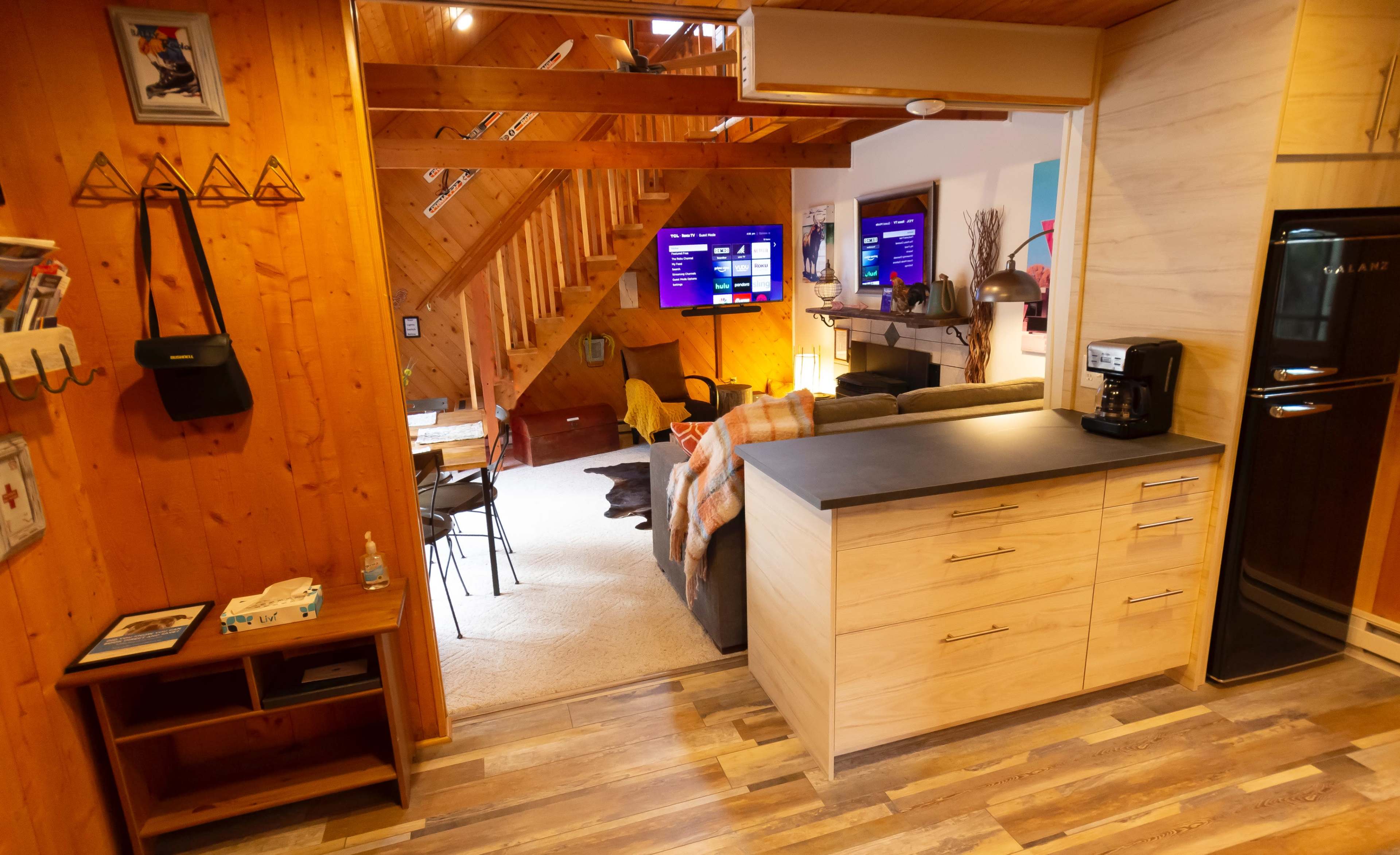 A cozy interior features a wooden stairway, two wall-mounted TVs, and a kitchen area with a coffee maker and smooth countertops.