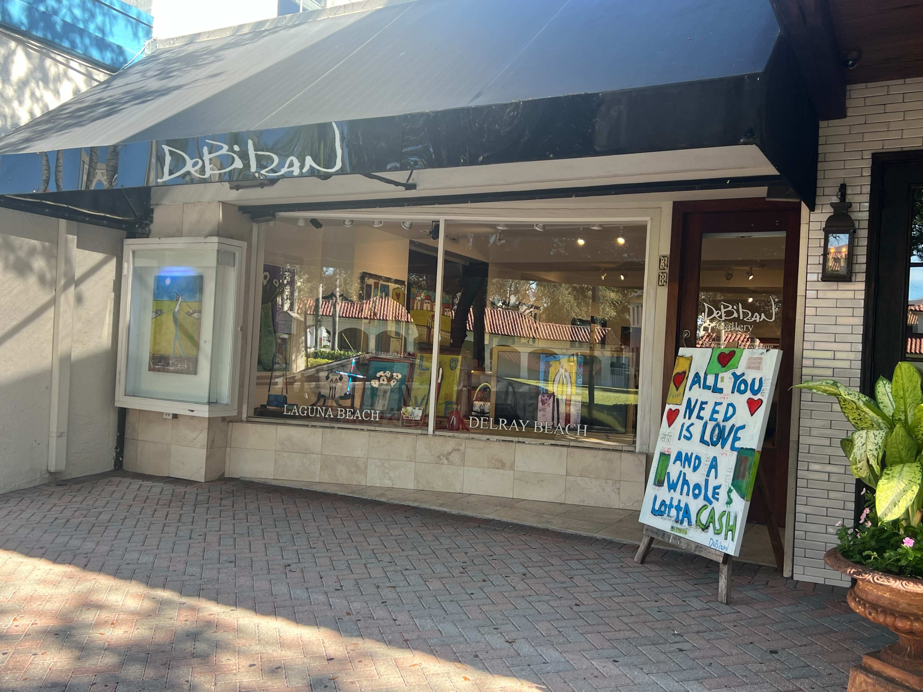 The image shows the exterior of an art gallery named "Debidan," featuring a storefront with large windows displaying colorful artwork and a sign stating, "All you need is love and a whole lotta blush."