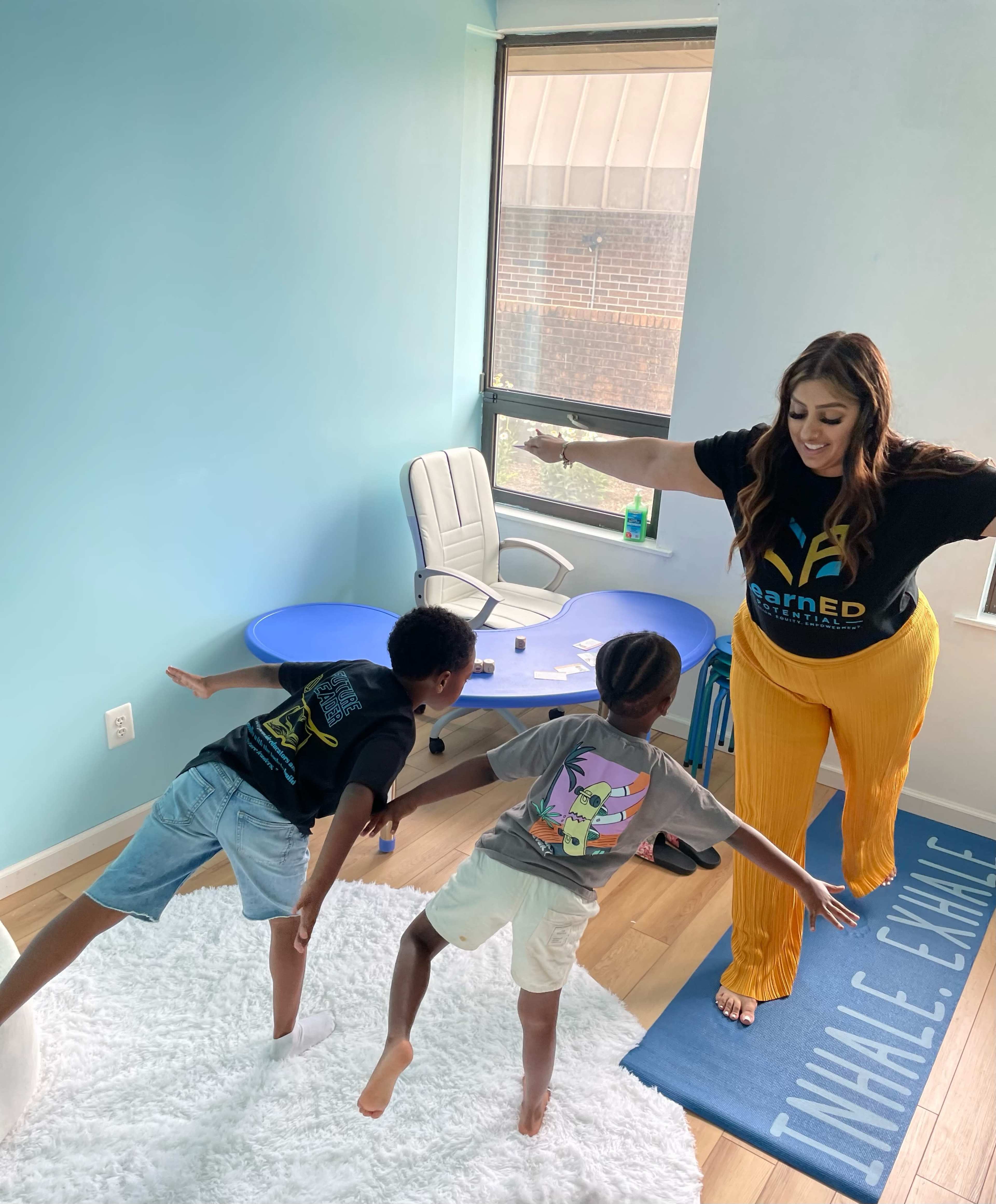 A woman demonstrates a yoga pose while two children mimic her in a brightly lit room with blue walls and a white rug.