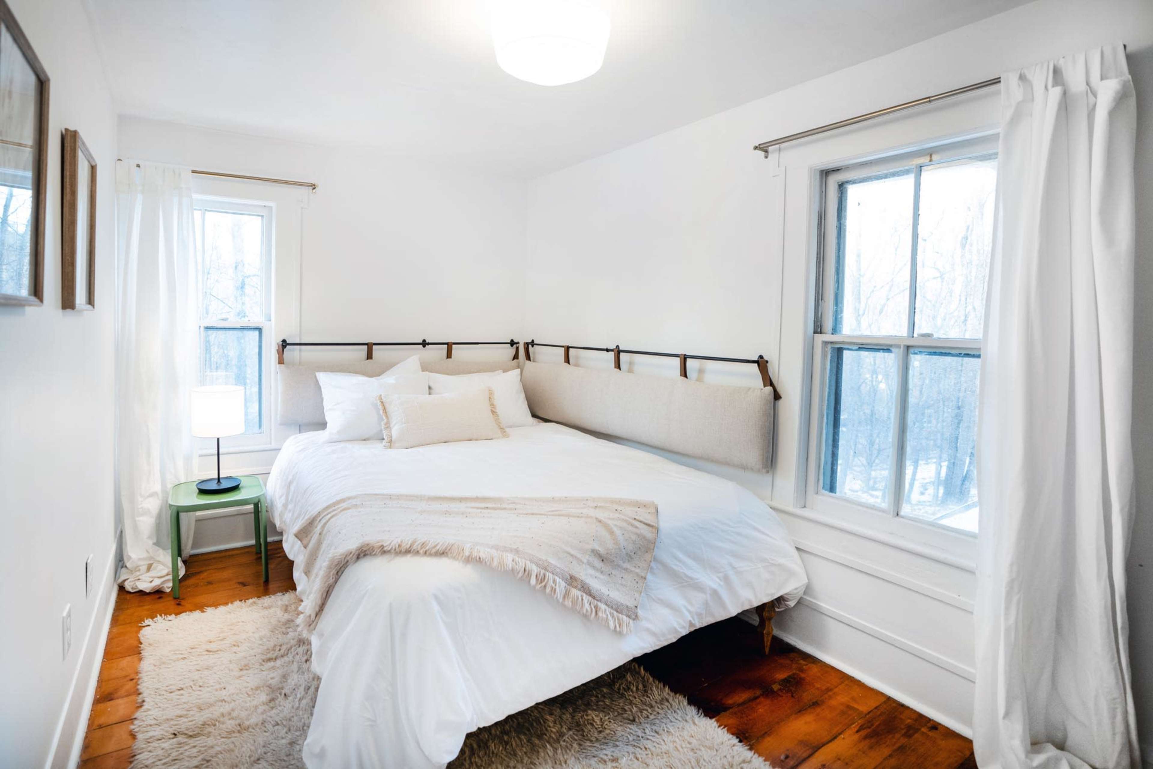 A bright bedroom features a large bed with a neutral headboard, two windows, a small green nightstand, and a lamp.