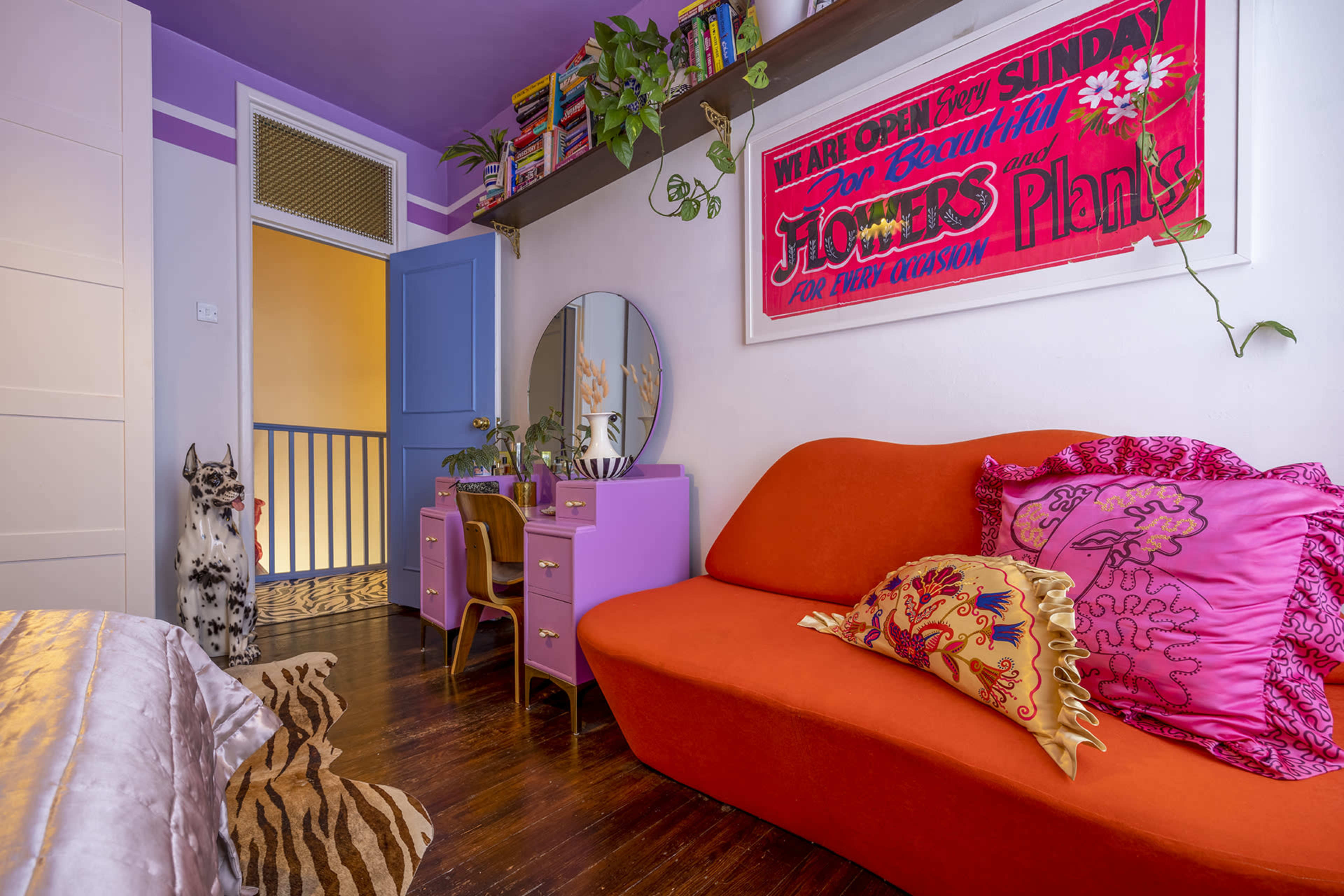A cozy room features an orange sofa, a purple dresser with a mirror, and a colorful wall hanging, alongside a staircase and a decorative dog statue.