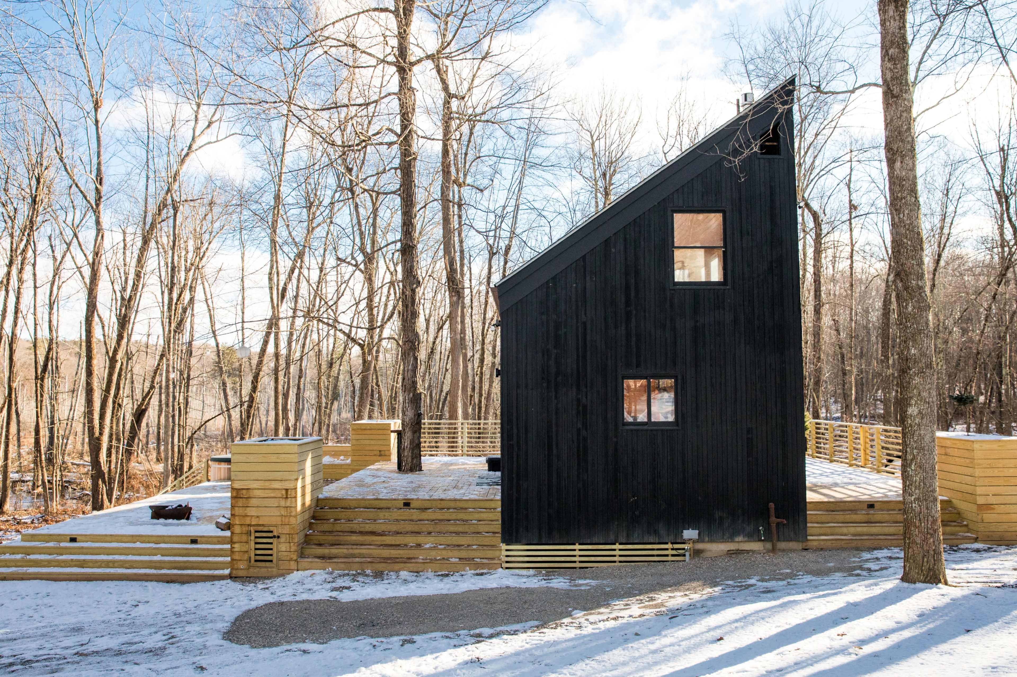 A modern black wooden house with a slanted roof, surrounded by snow and trees in a wooded area.