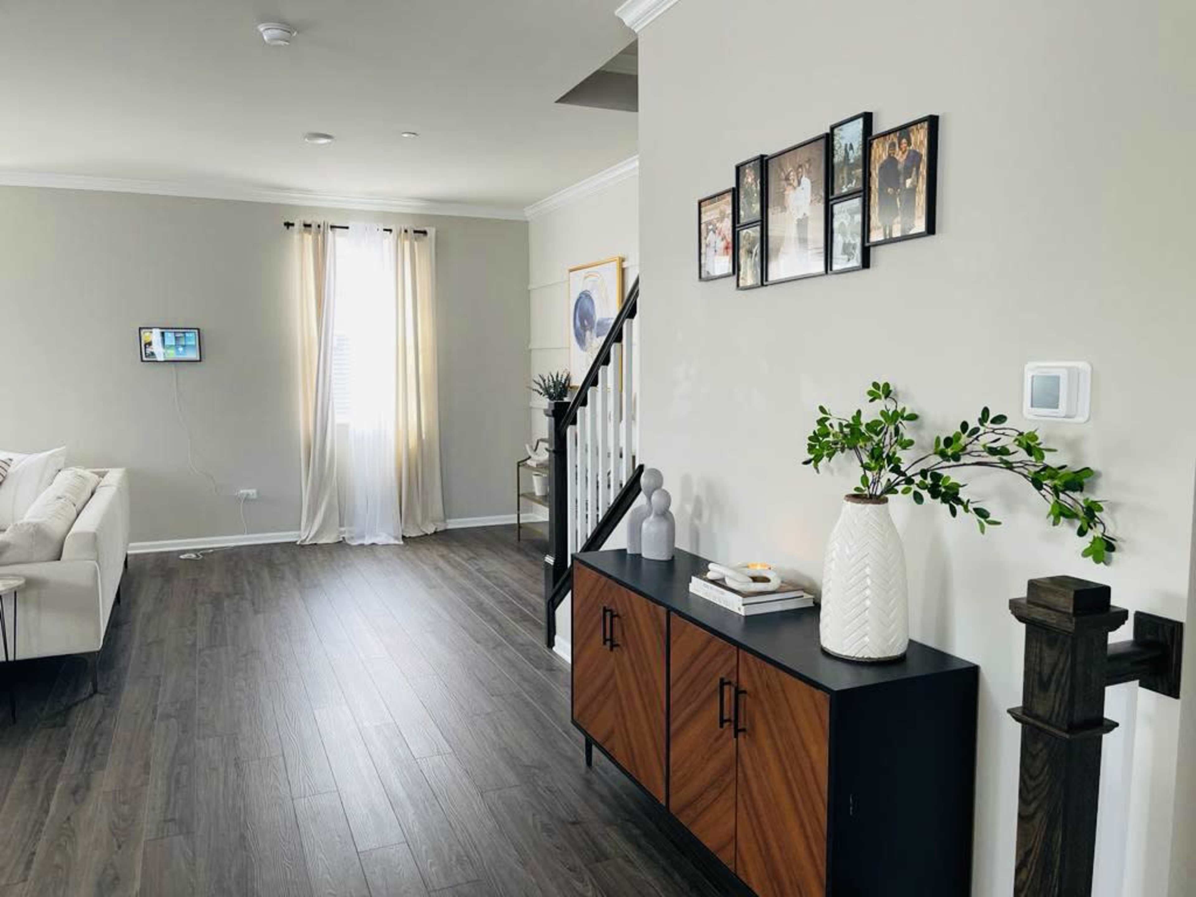 The image shows a modern living room entryway with a wooden console table, framed photographs on the wall, and large curtains by a window.