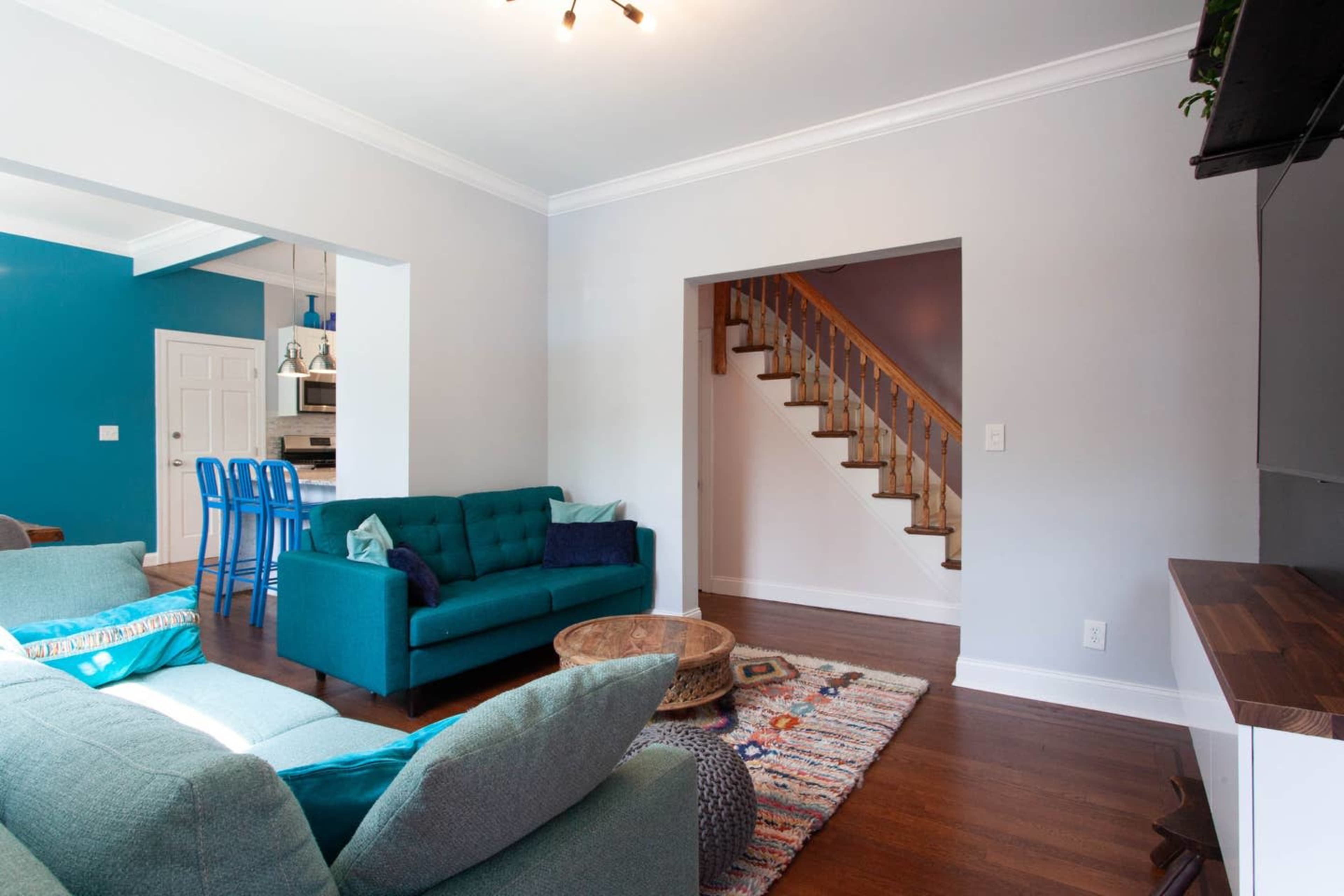 The image shows a living room with a teal couch, a wooden coffee table, and a staircase leading to an upper level.