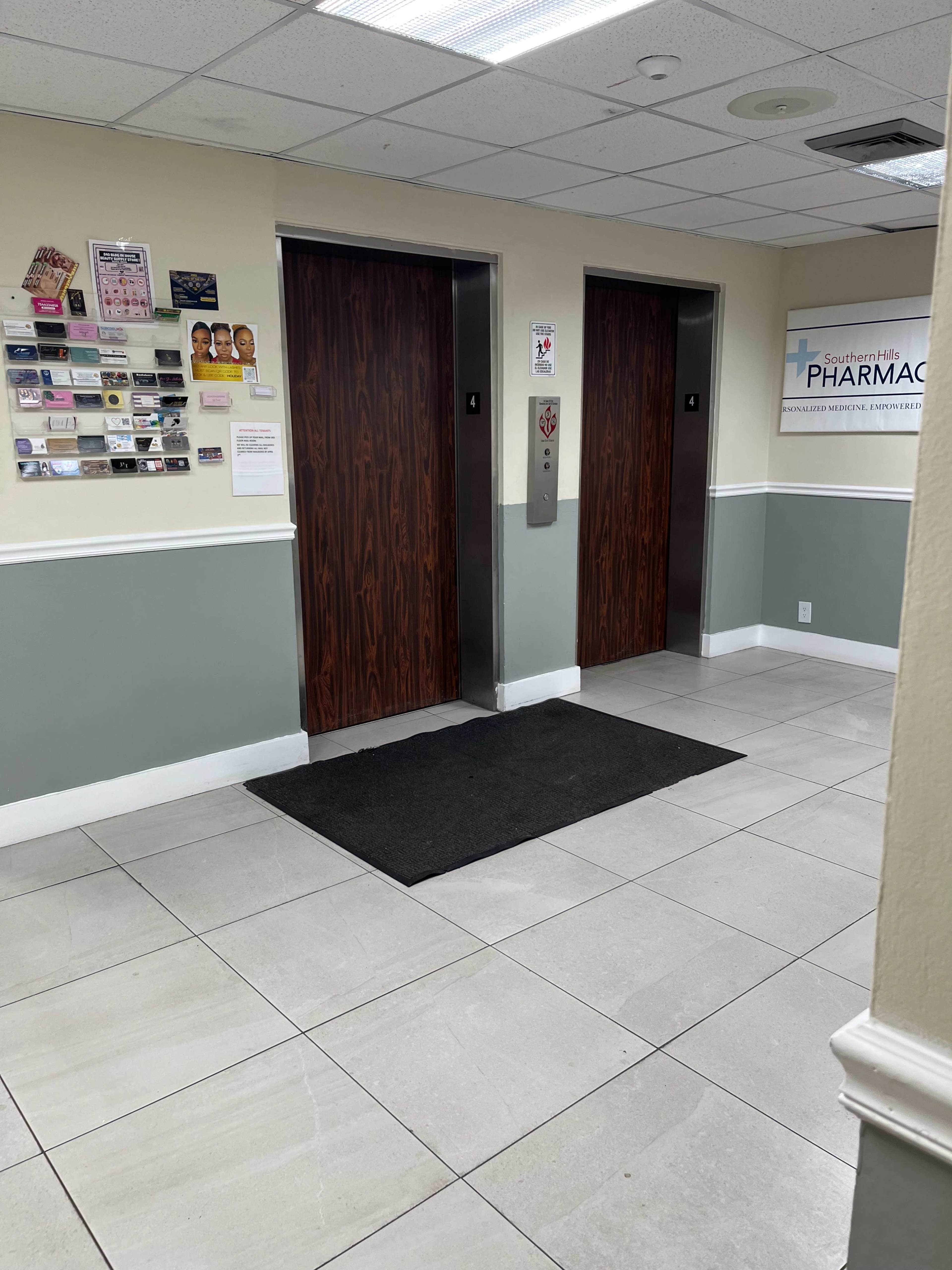 A hallway with two elevator doors, a welcome mat in front, and a wall displaying various cards and signage.