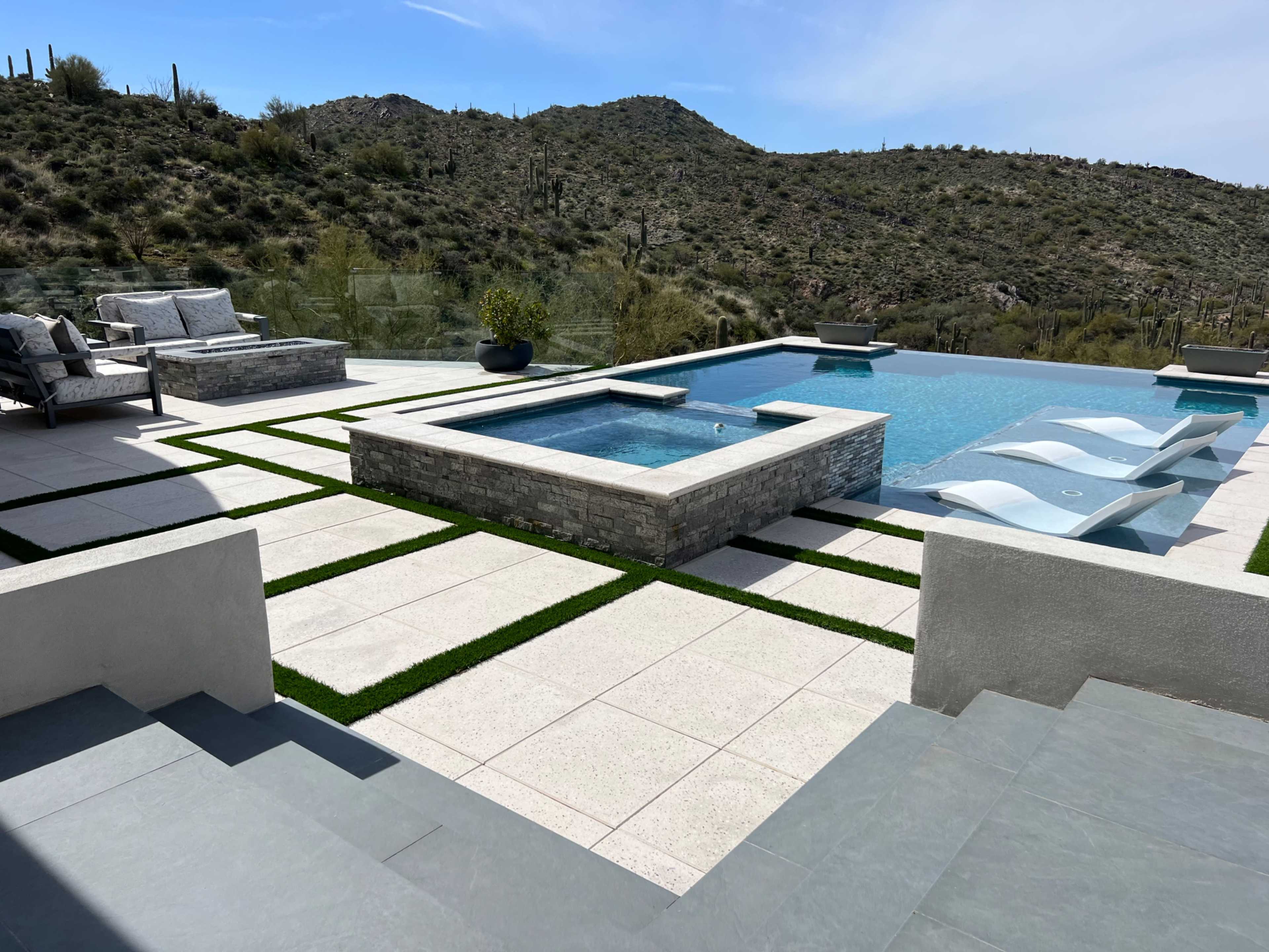 The image shows a modern outdoor pool area with a water feature, lounge chairs, and a view of cacti-covered hills in the background.