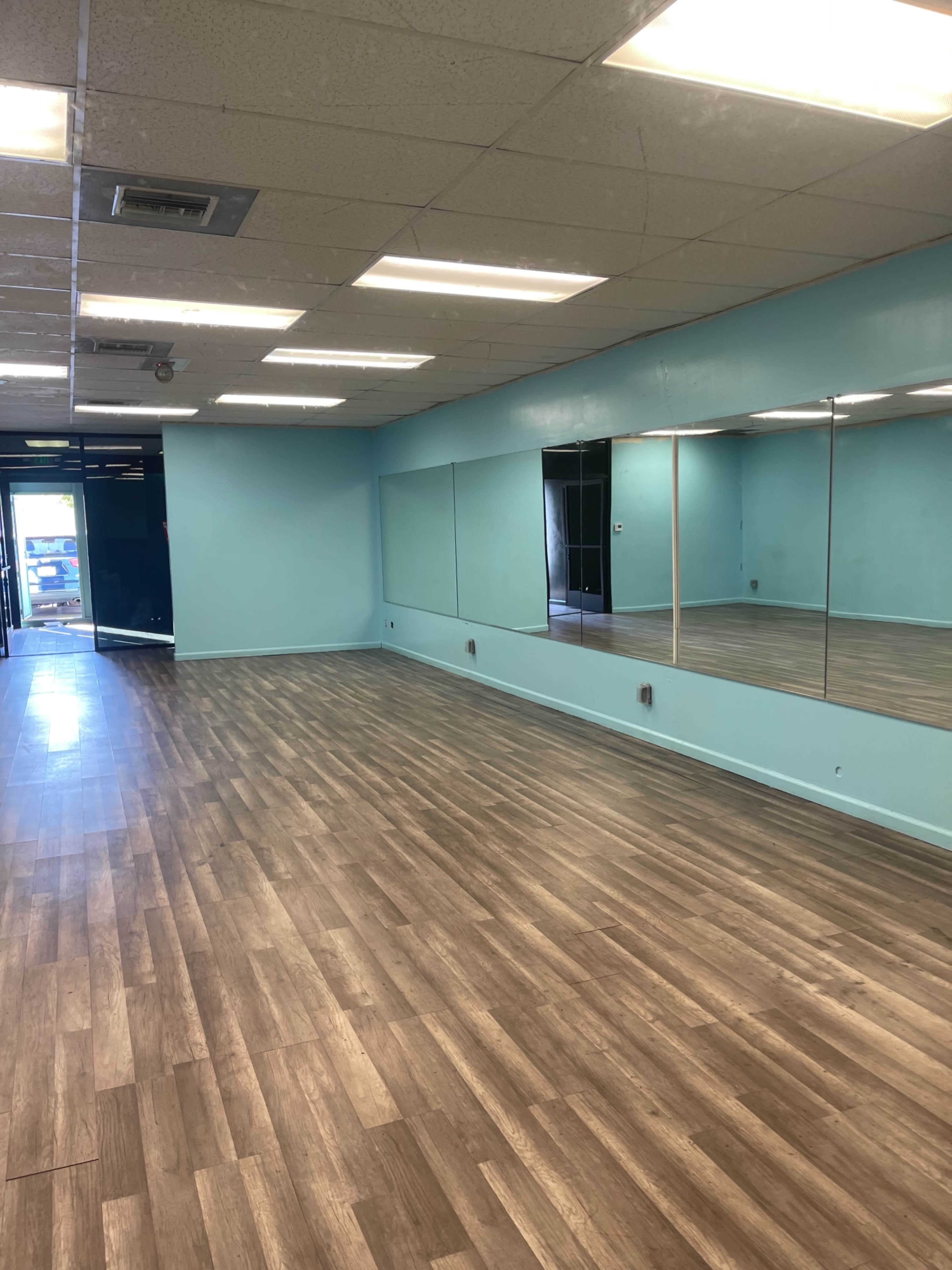 A spacious, empty room with a light blue wall and large mirrors, featuring a wooden floor and overhead fluorescent lighting.