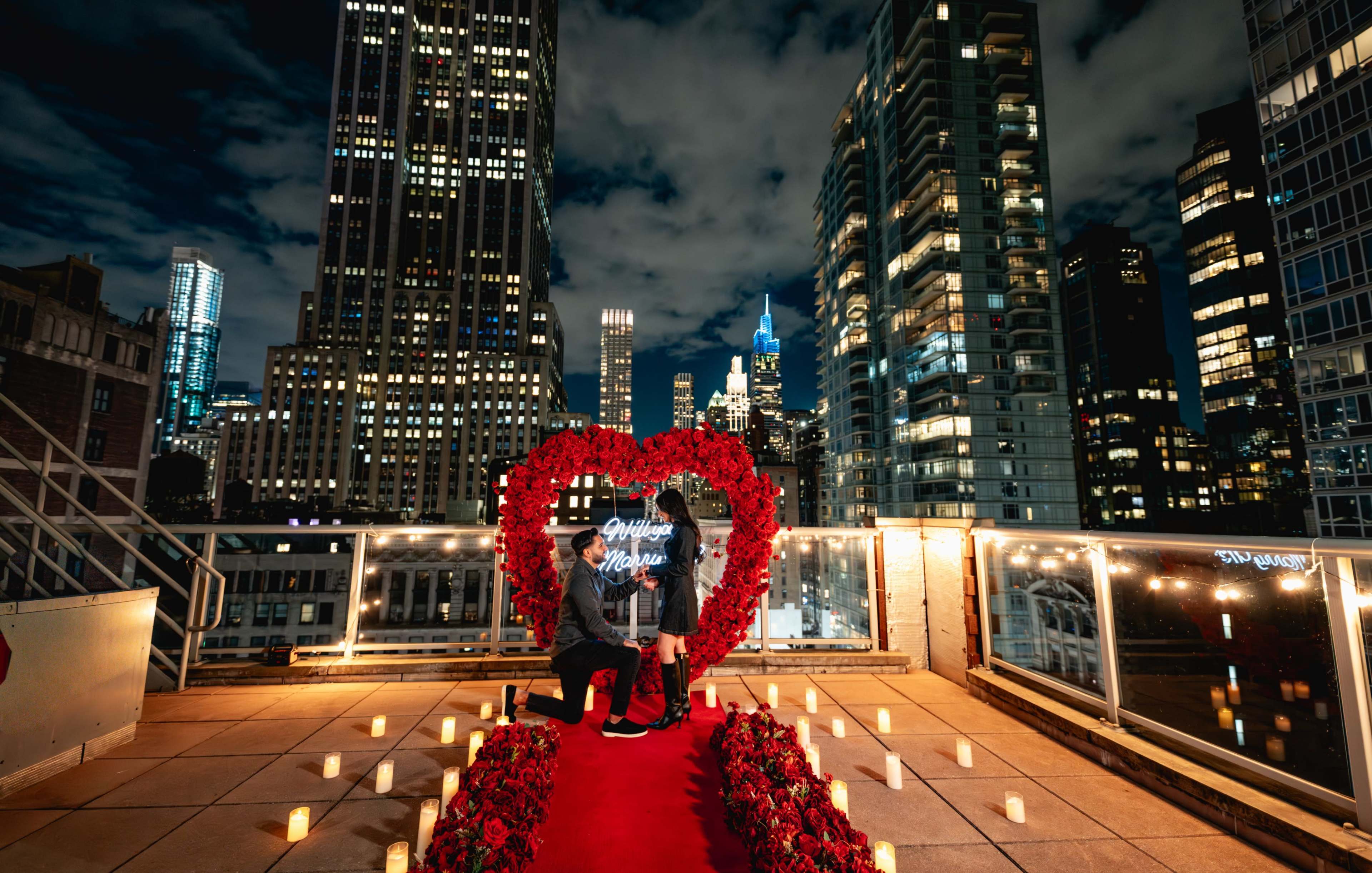 A man is proposing to a woman on a rooftop decorated with a heart-shaped arrangement of red roses and candles, with a city skyline in the background at night.