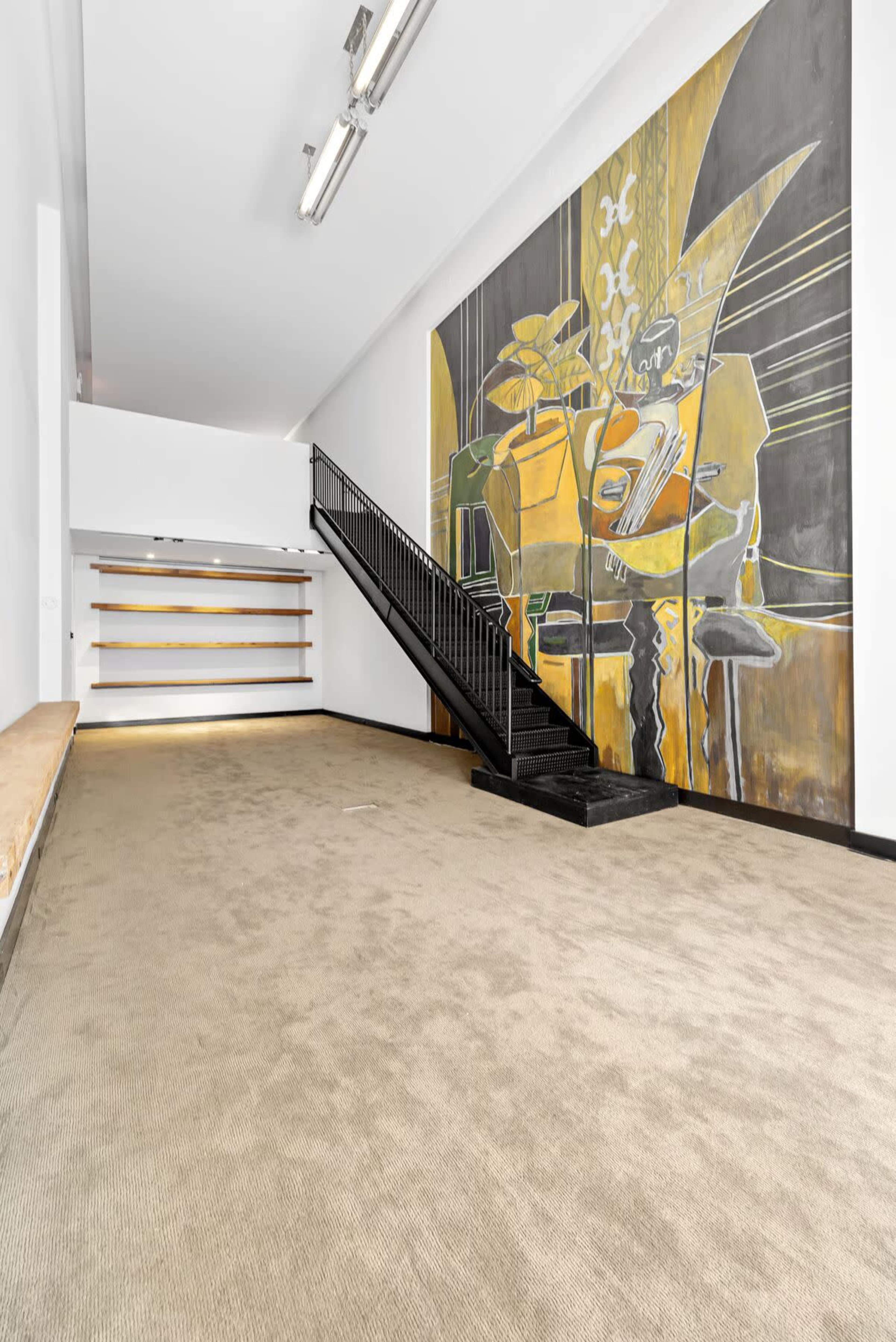 The interior of a spacious room features a staircase on one side and a large mural depicting a figure, along with empty shelves against the wall.