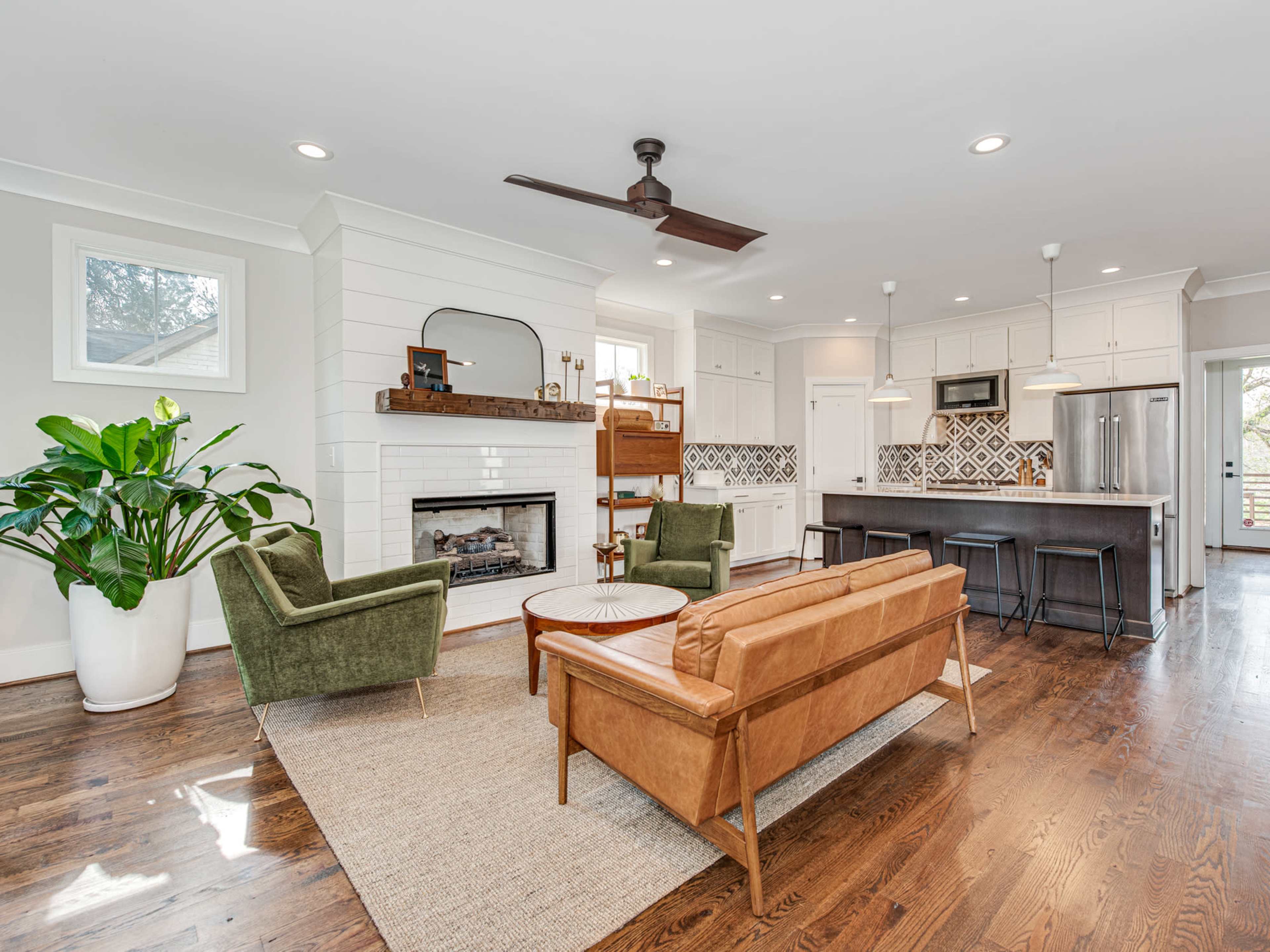 A modern living room features a brown leather sofa, green armchairs, a fireplace, and a kitchen area with patterned tiles and bar stools.