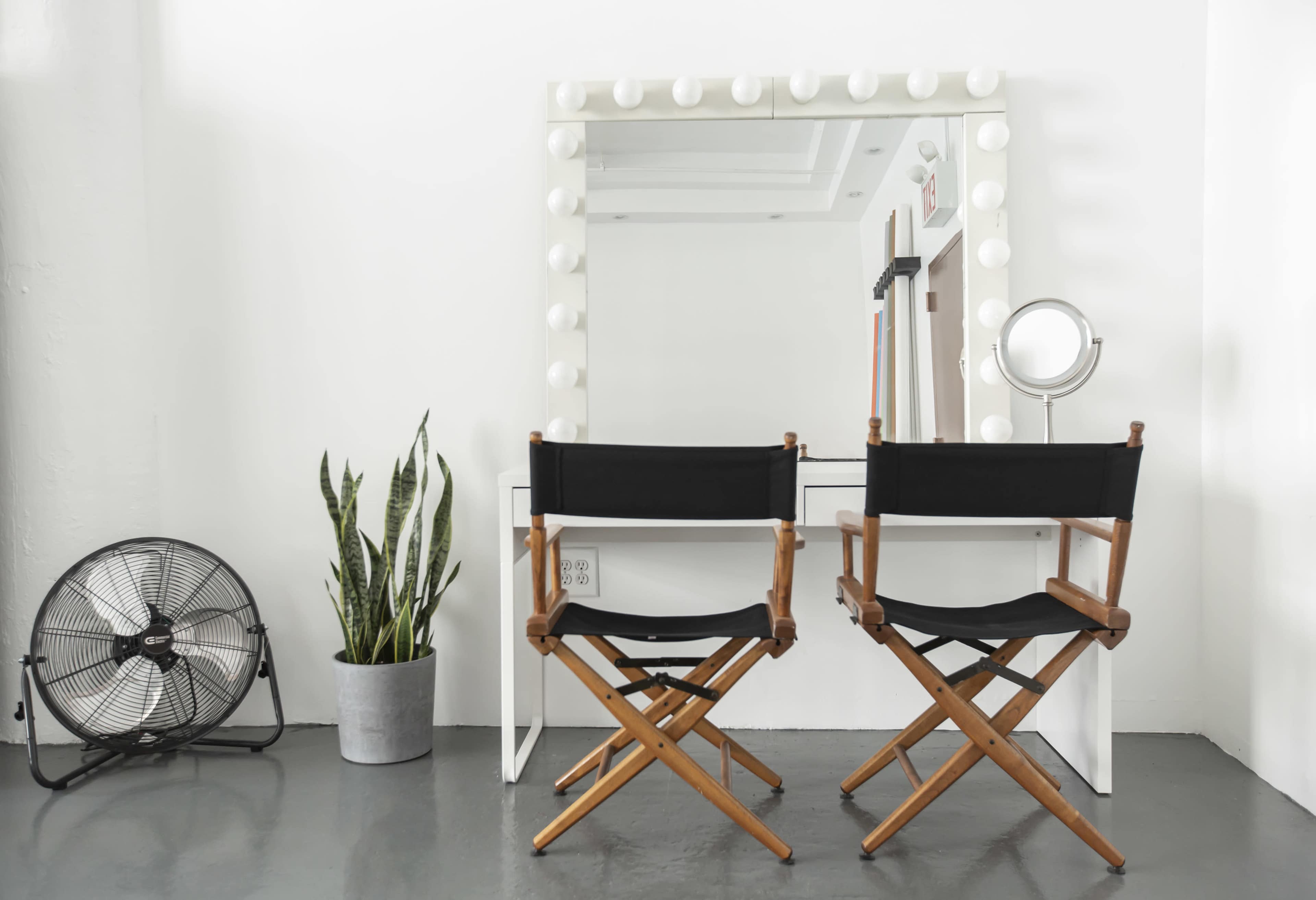 The image shows a minimalist dressing area with two wooden director's chairs facing a large illuminated mirror, a small round mirror, a potted plant, and a fan nearby.