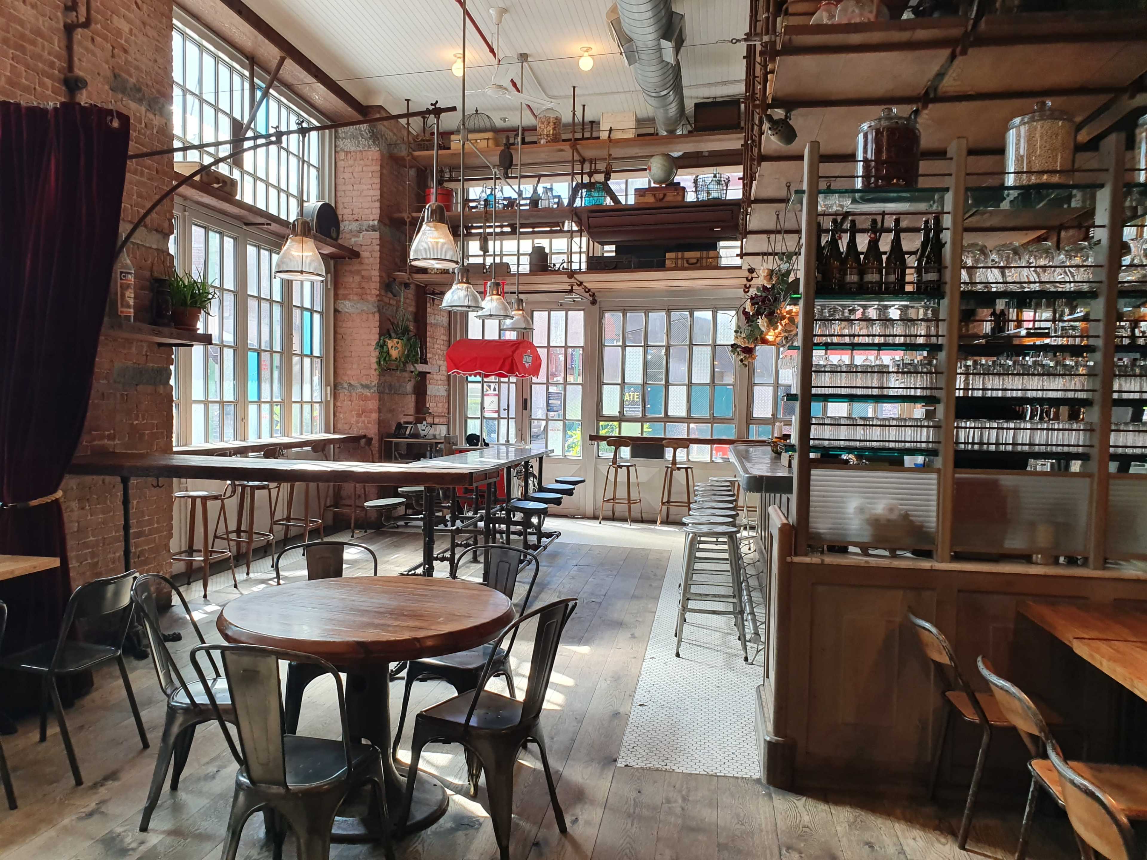 A spacious interior of a café or restaurant featuring wooden tables, industrial-style chairs, large windows, and a well-organized bar area.