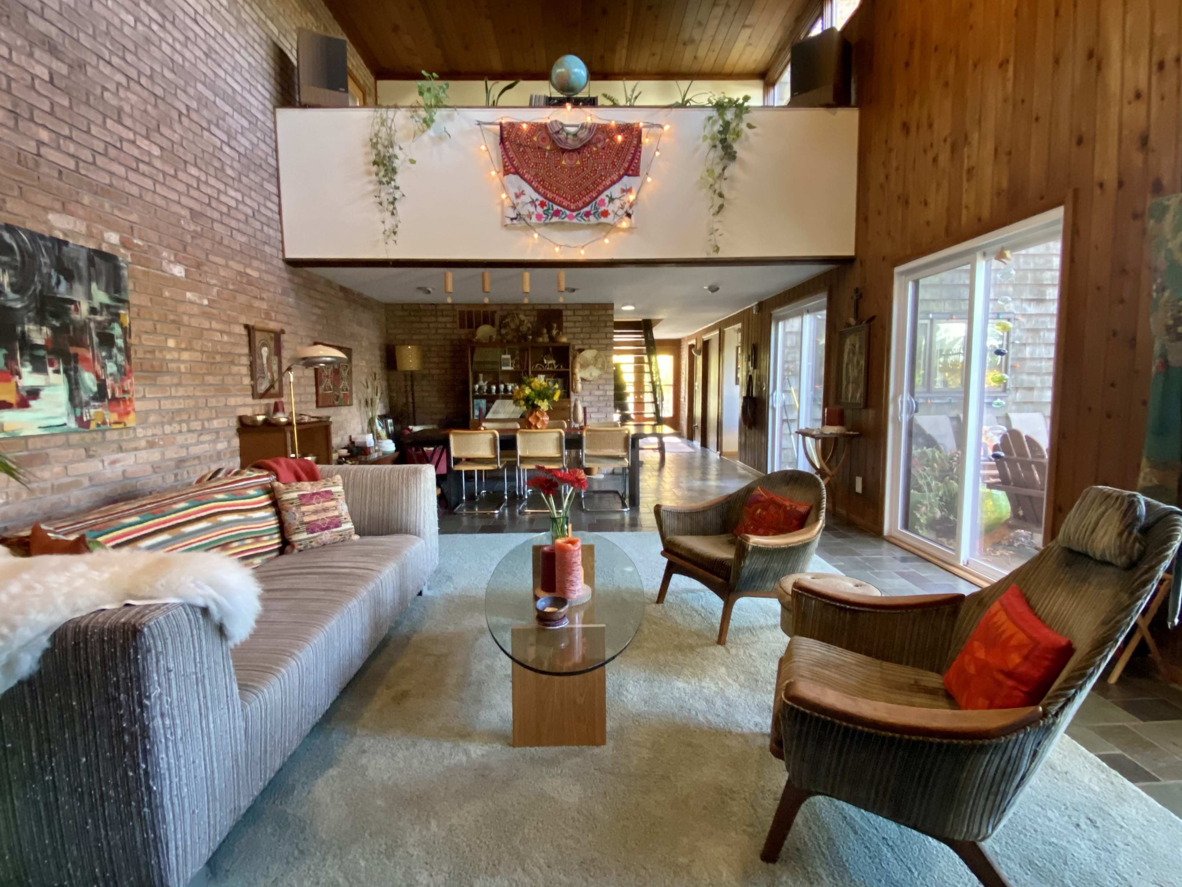 The image shows a modern living room with a sofa, armchairs, a glass coffee table, and large windows that open to a hallway and kitchen area.