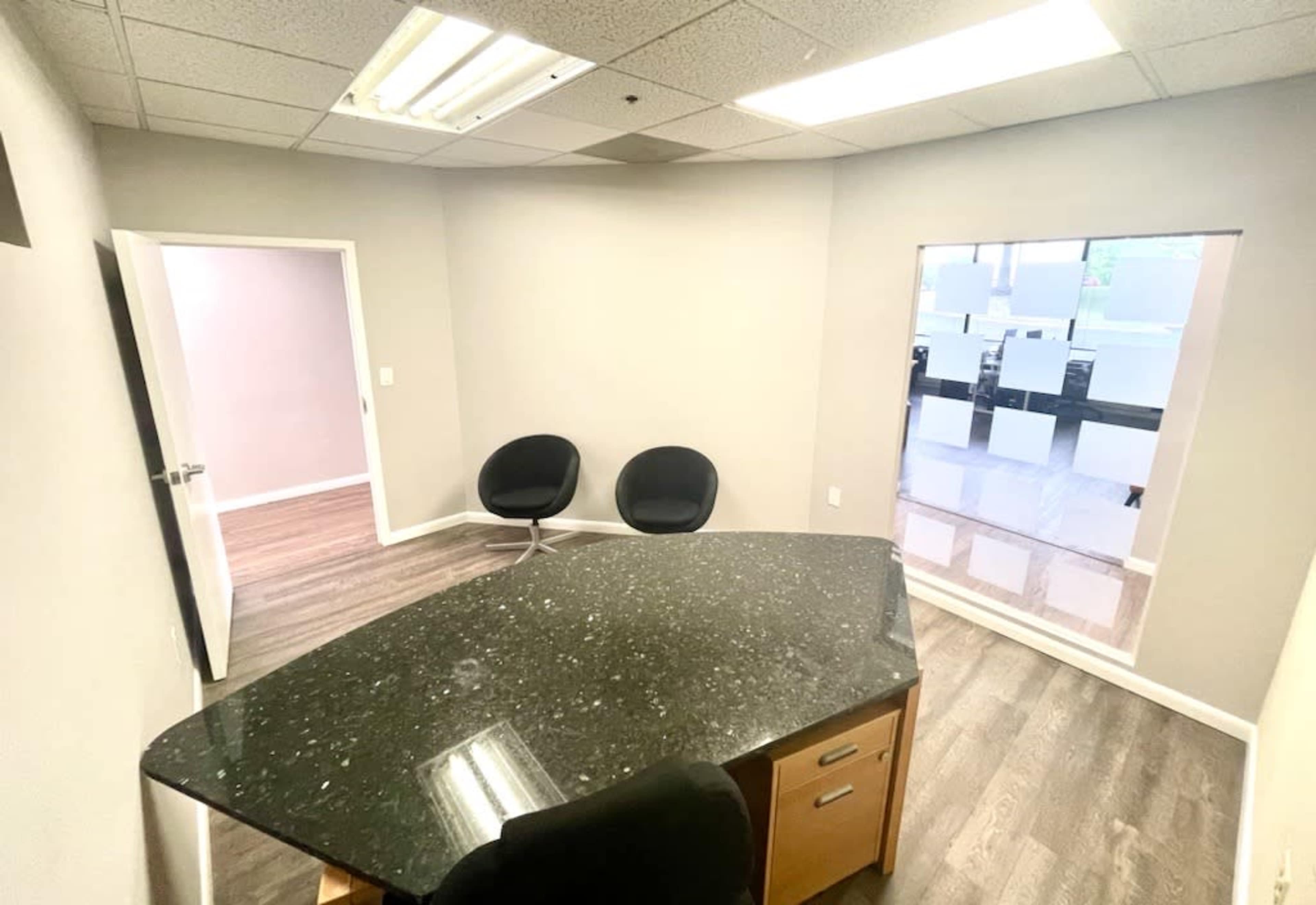 A small office space features a dark granite desk, two black chairs, and a large window with frosted glass.