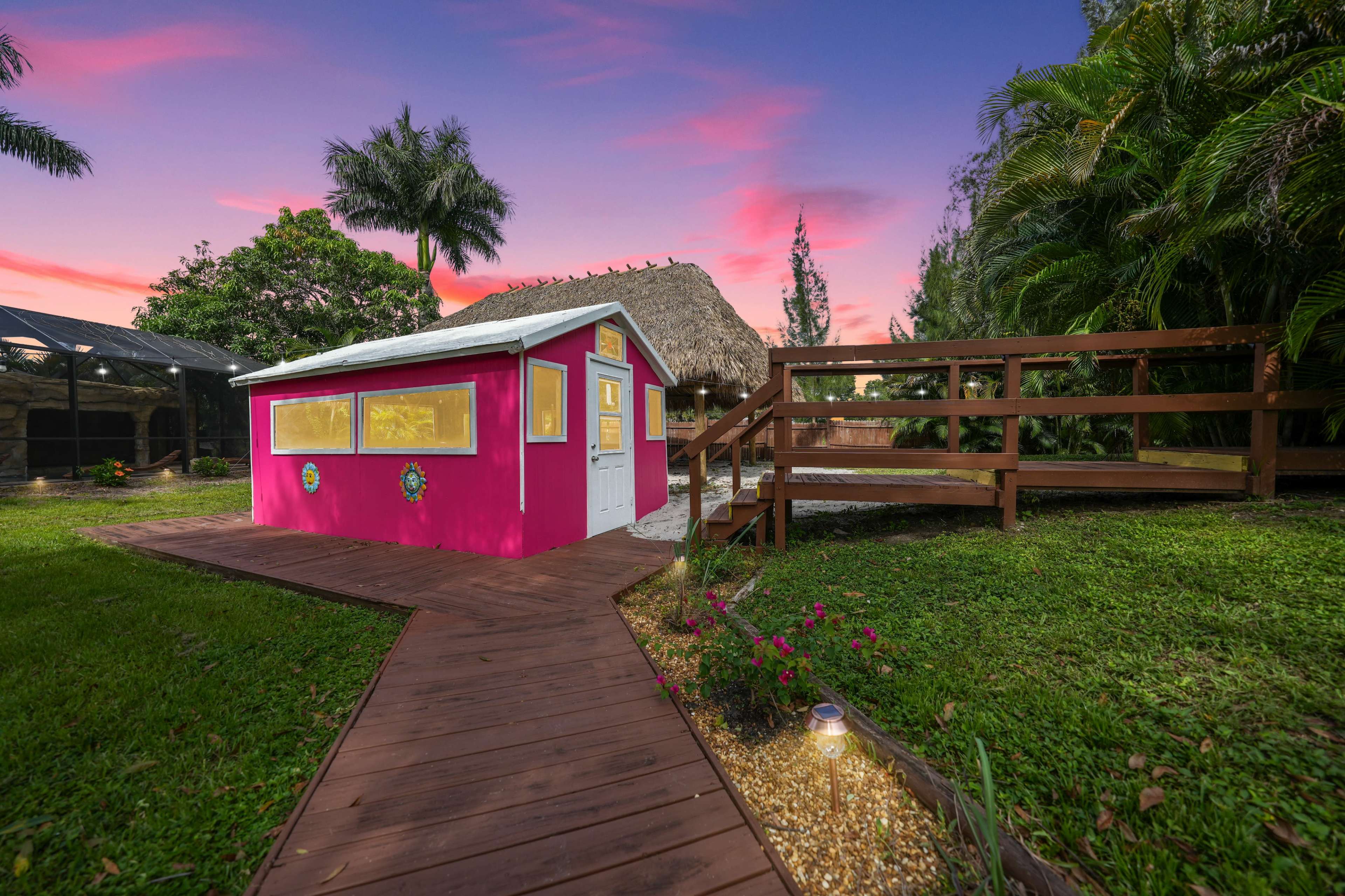 A colorful pink shed with yellow windows situated next to a thatched-roof structure, surrounded by lush greenery and a wooden walkway leading to a raised deck area at sunset.