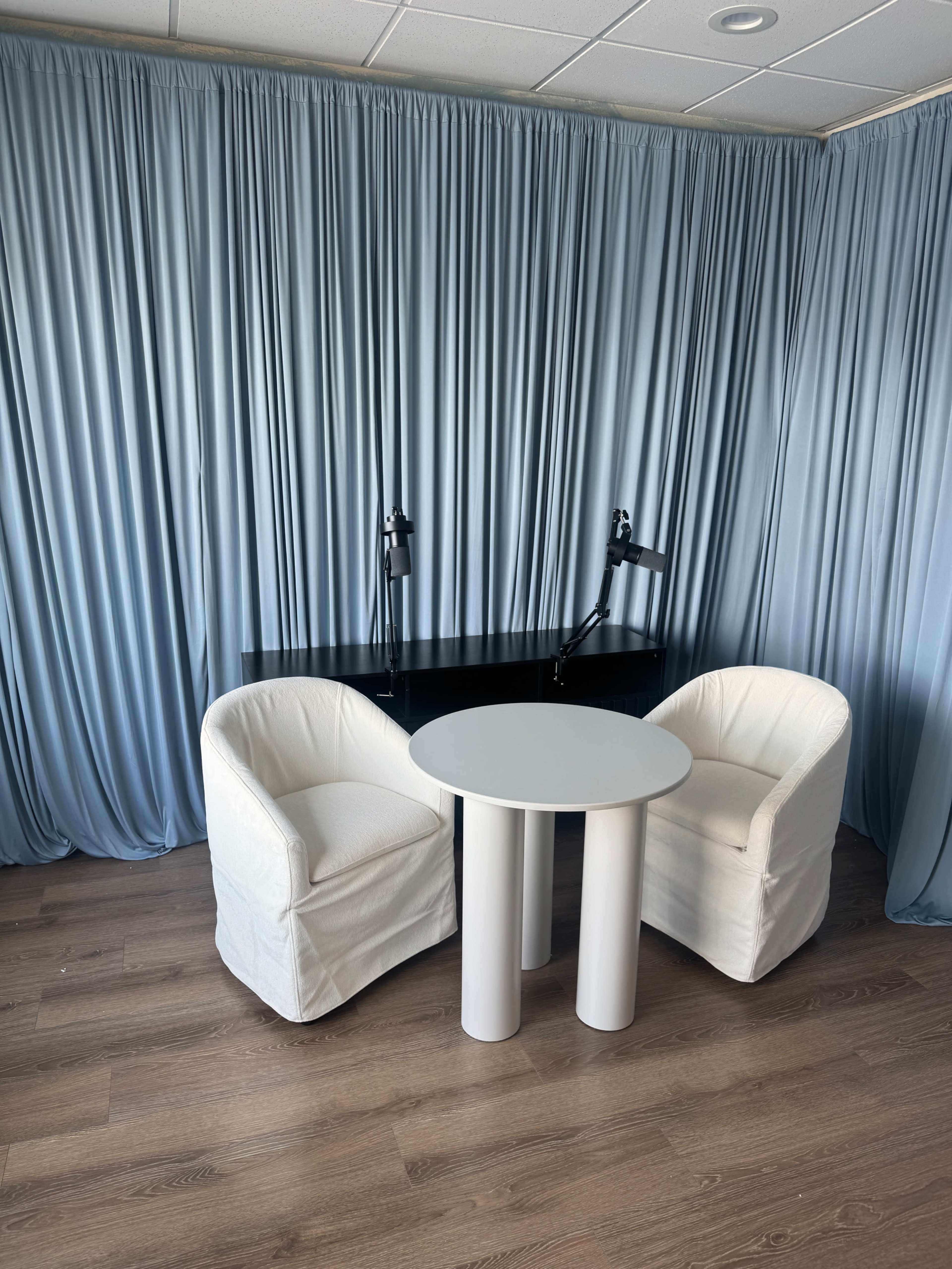 The image shows a small seating area with two white armchairs and a round table in front of a backdrop of blue curtains.