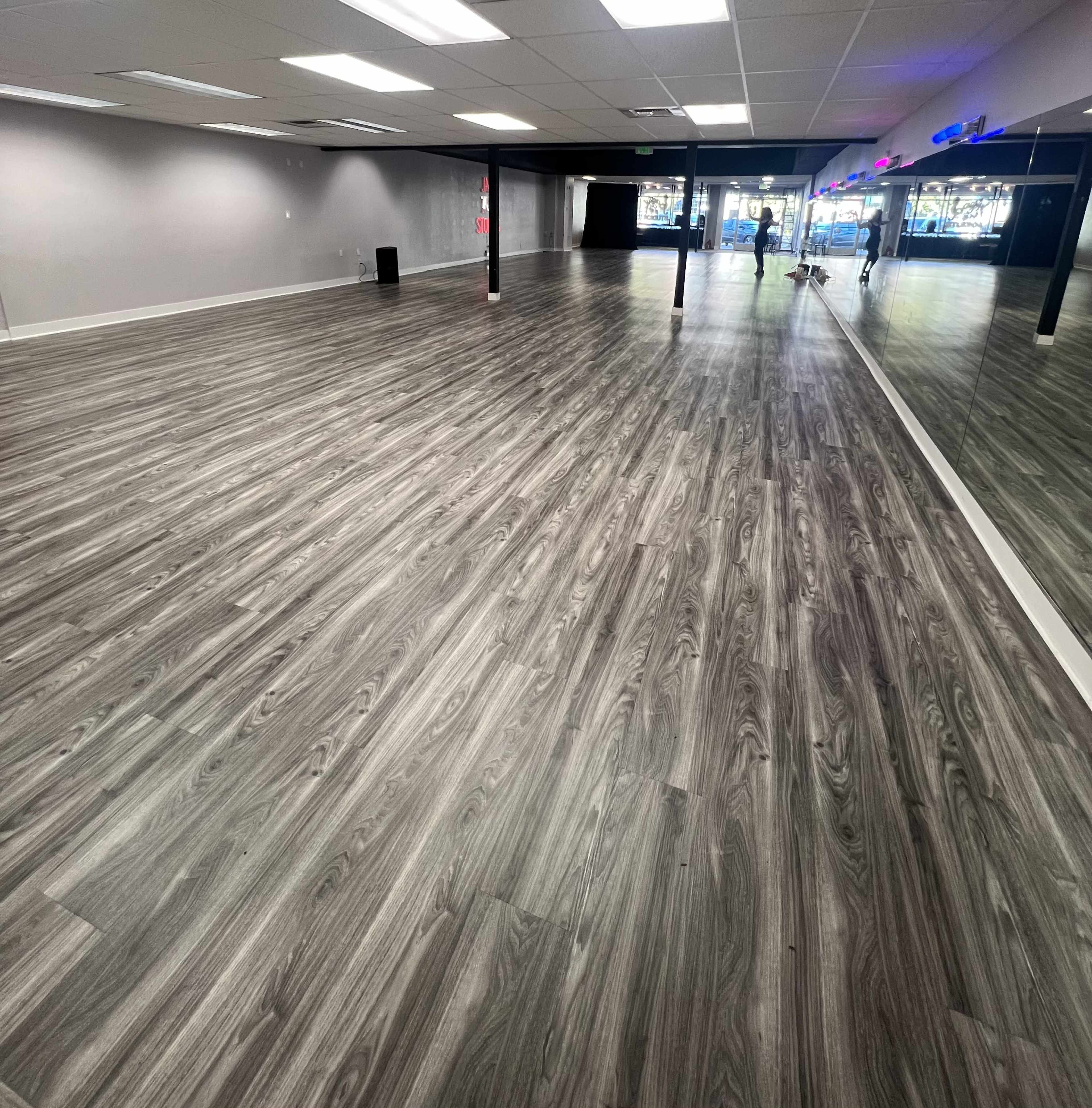 The image shows an empty indoor space with gray wooden flooring and mirrors lining one wall.
