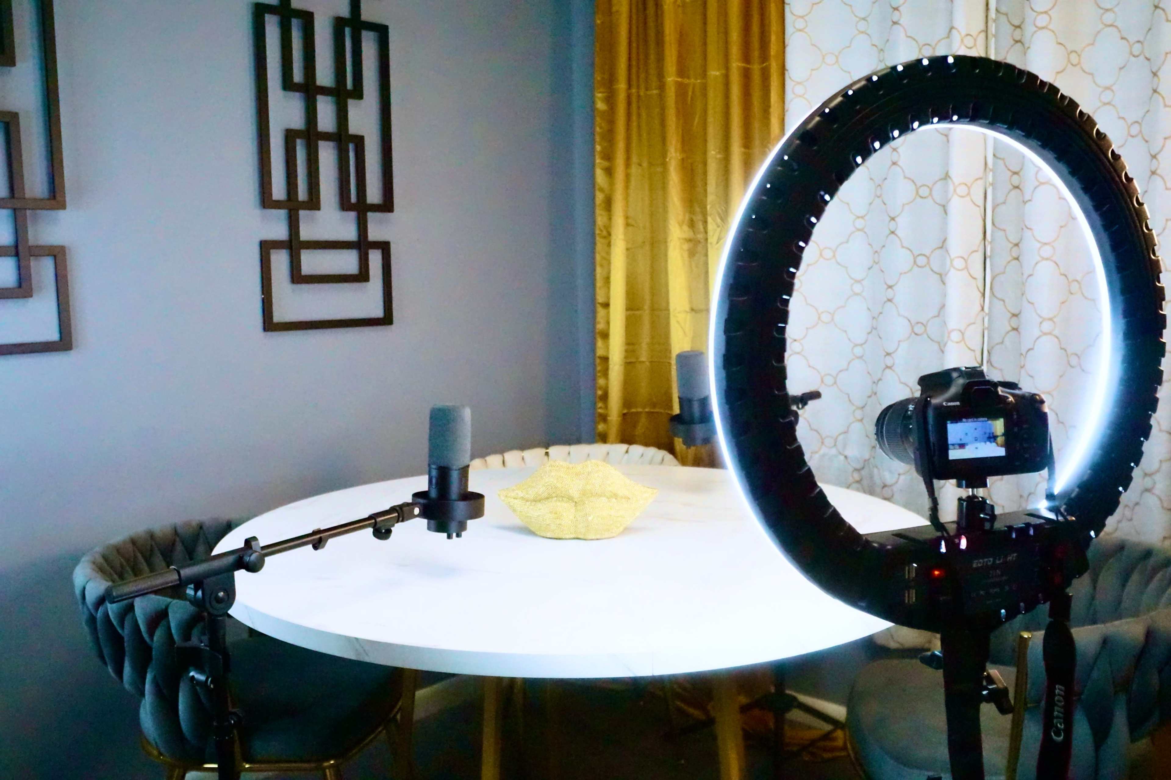 A round marble table is set with two microphones and a camera mounted on a tripod, illuminated by a ring light in a well-decorated room.