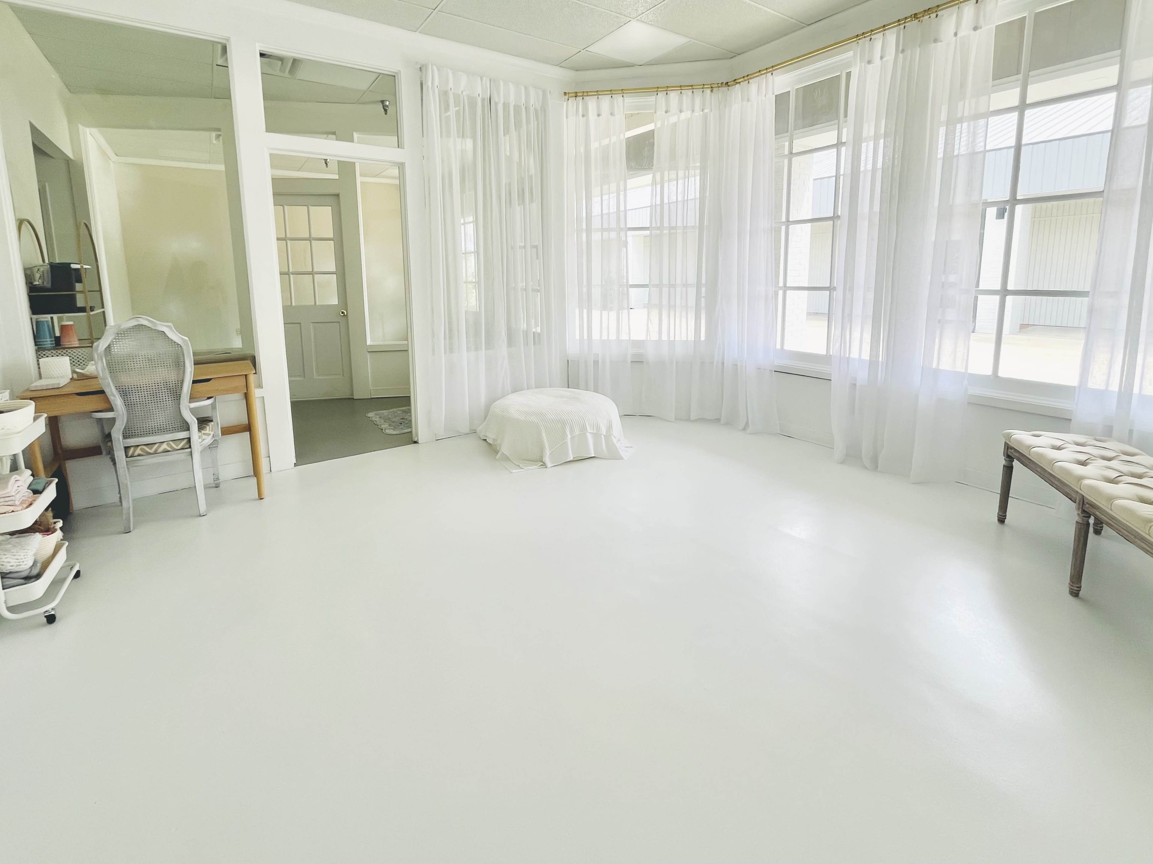 A bright, airy room with white walls and flooring, featuring large windows draped with sheer curtains and minimal furniture, including a desk and a cushioned bench.