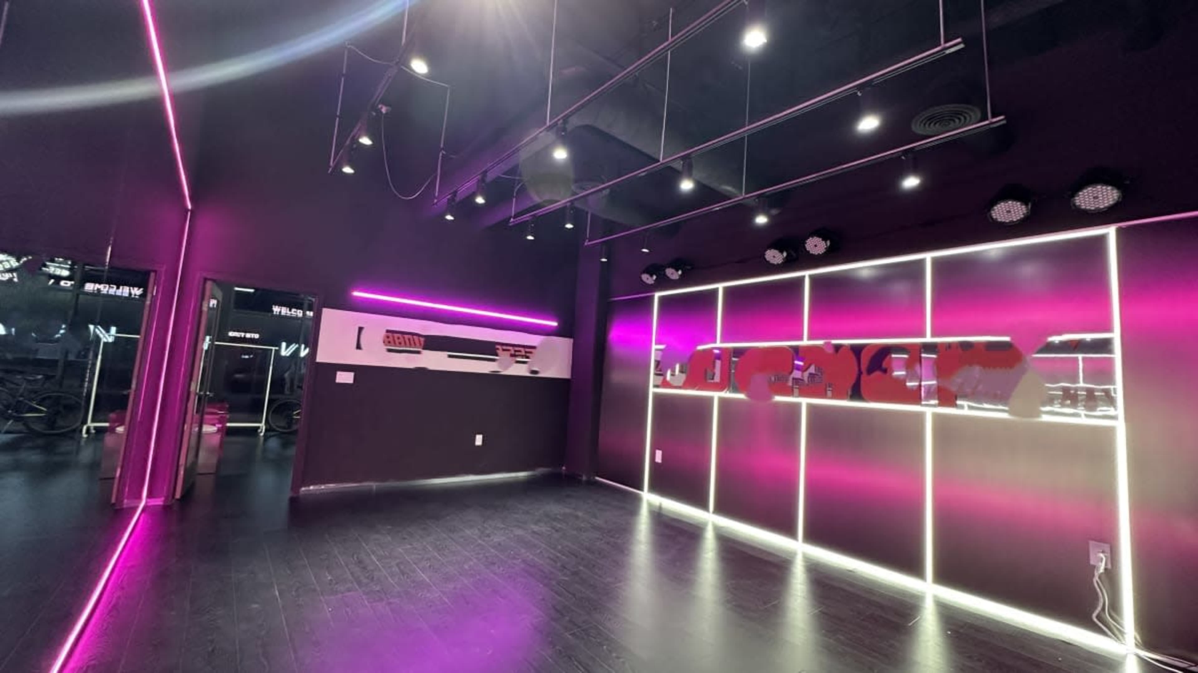 The image shows a modern fitness studio with black walls, neon pink lighting, and a mirrored section.