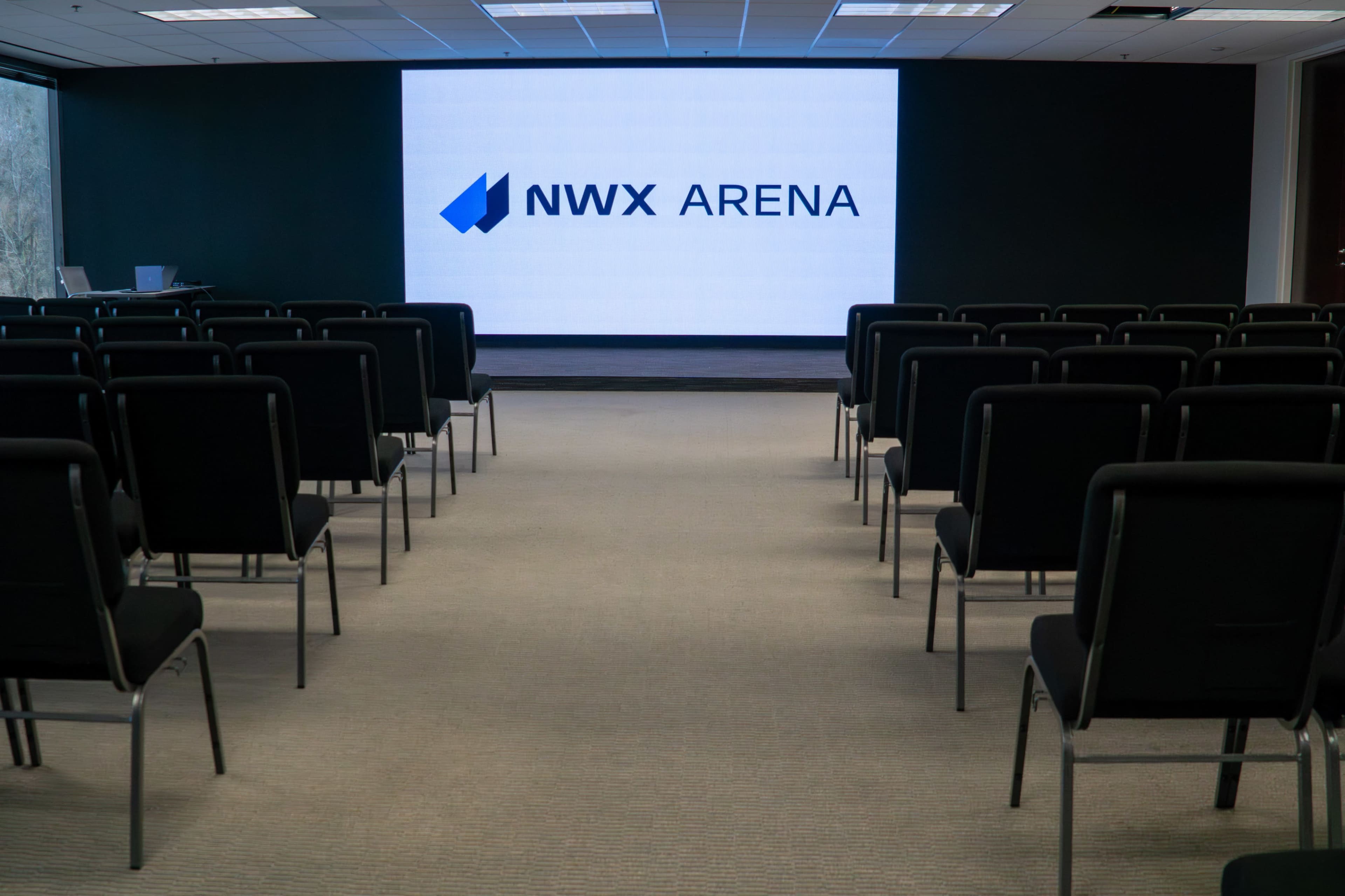 The image shows a conference room with rows of black chairs facing a large screen displaying the logo "NWX ARENA" on a white background.