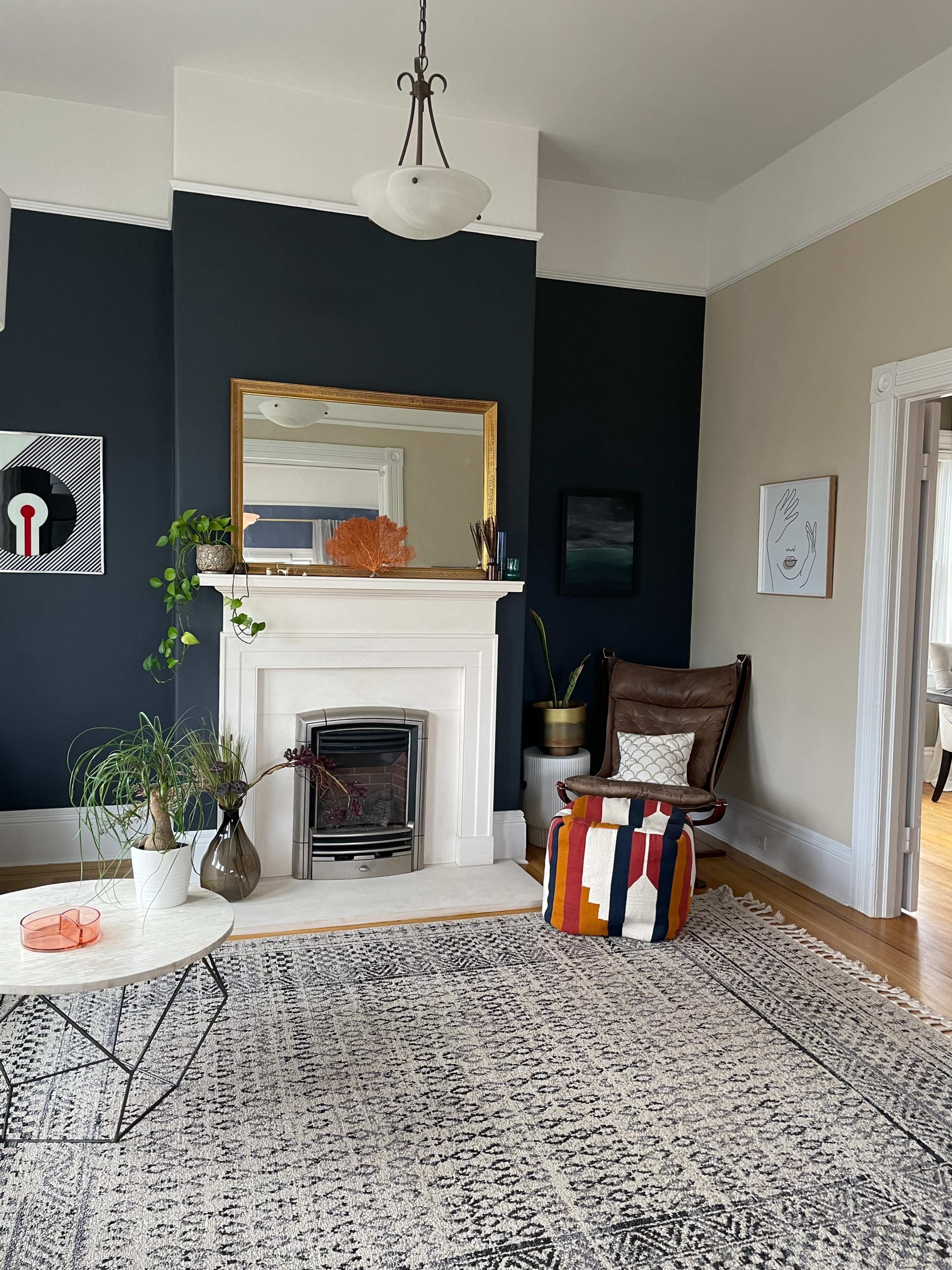 The living room features a fireplace with a mirror above it, a patterned rug, a modern chair, and a variety of potted plants against a dark wall.