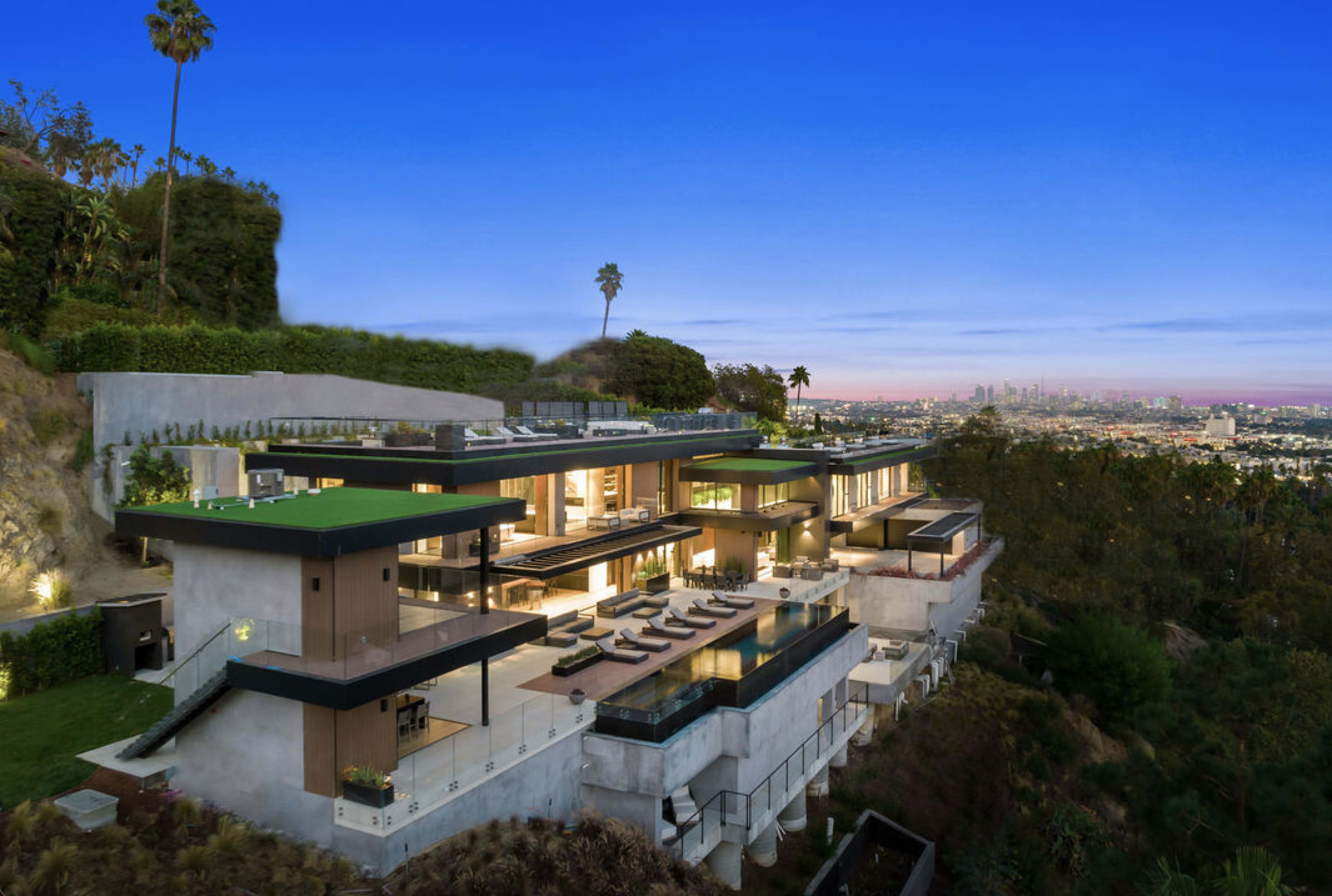 A modern multi-level house features large glass panels and a swimming pool, set on a hillside with a view of the city skyline.