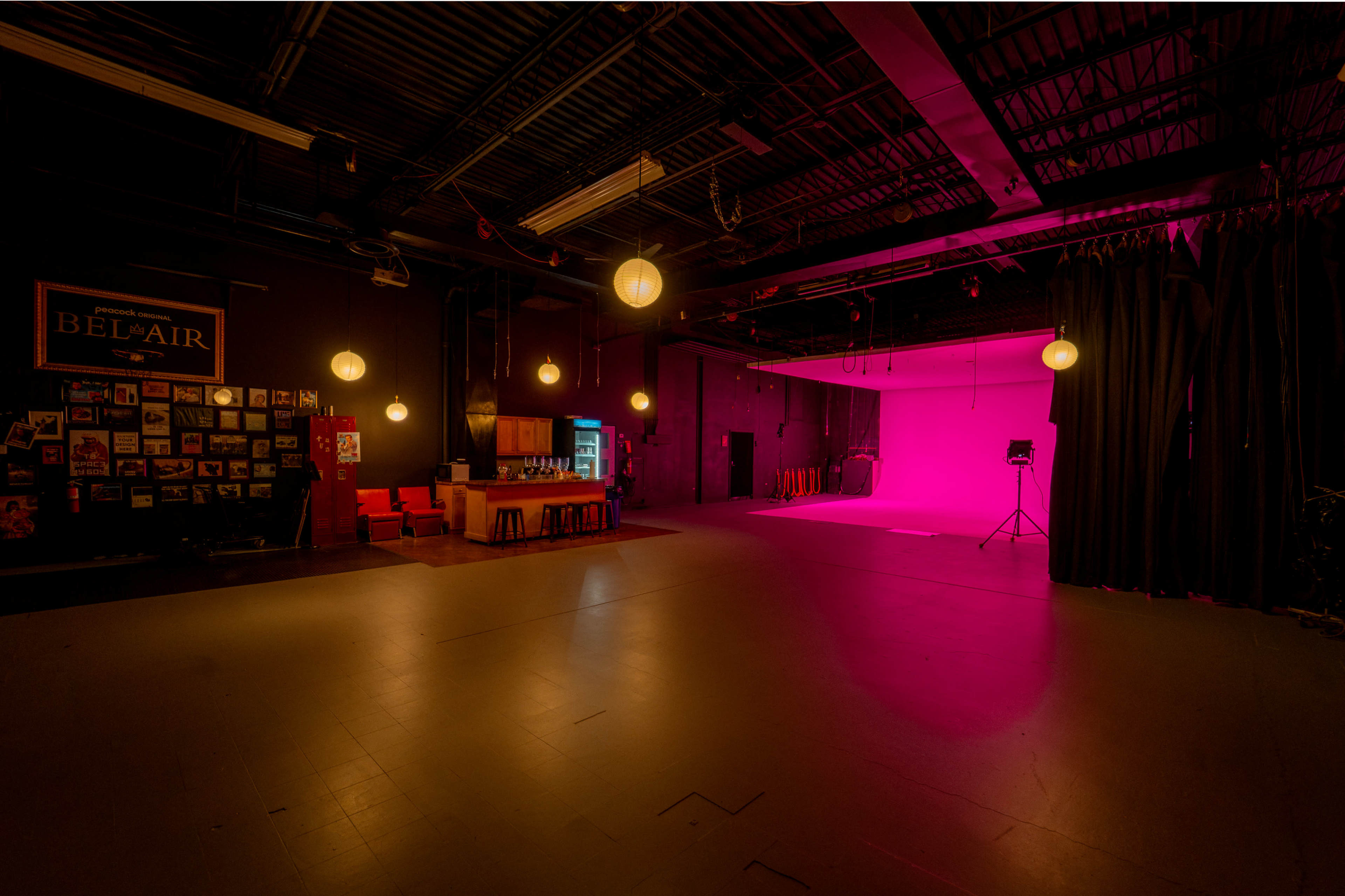 The image shows a spacious studio with a pink backdrop, illuminated by hanging lights, and features a seating area with wooden furniture and a wall of photographs.