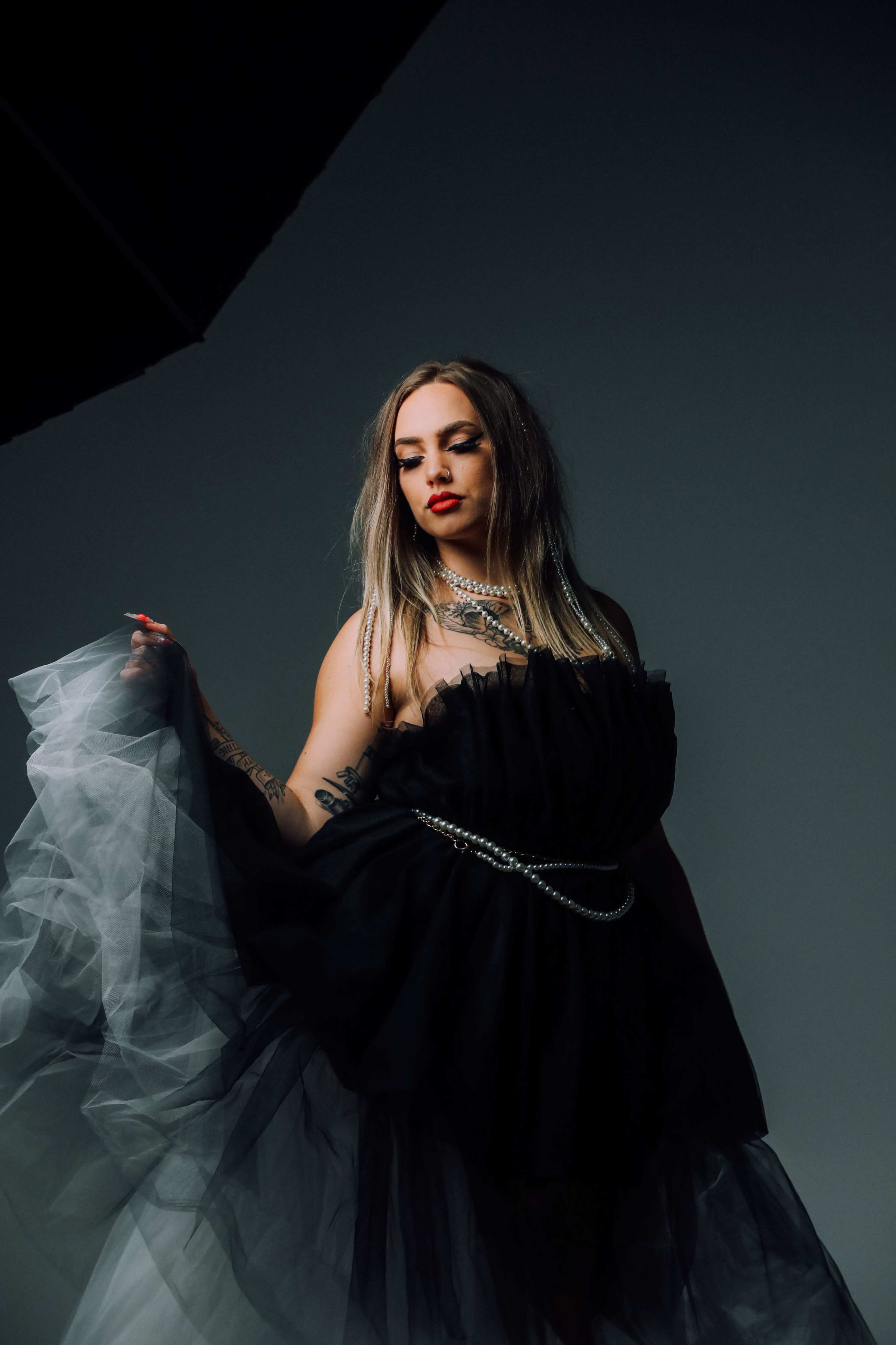 A woman poses in a black tulle dress, holding the fabric while wearing layered necklaces and bold red lipstick.