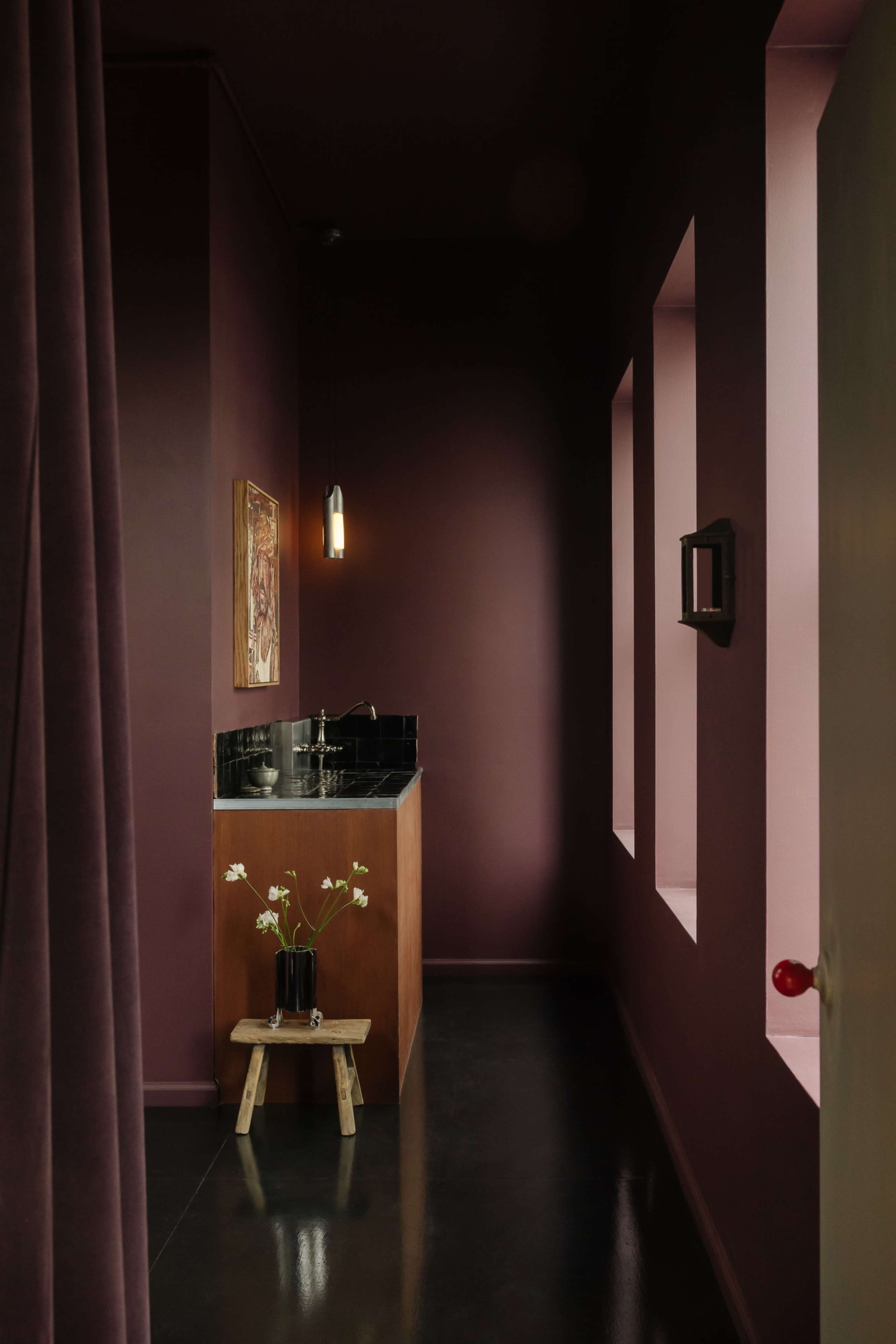 The image shows a hallway leading to a small kitchenette with a dark countertop against a deep maroon wall, lit by a hanging light and featuring a vase of flowers on a wooden stool.