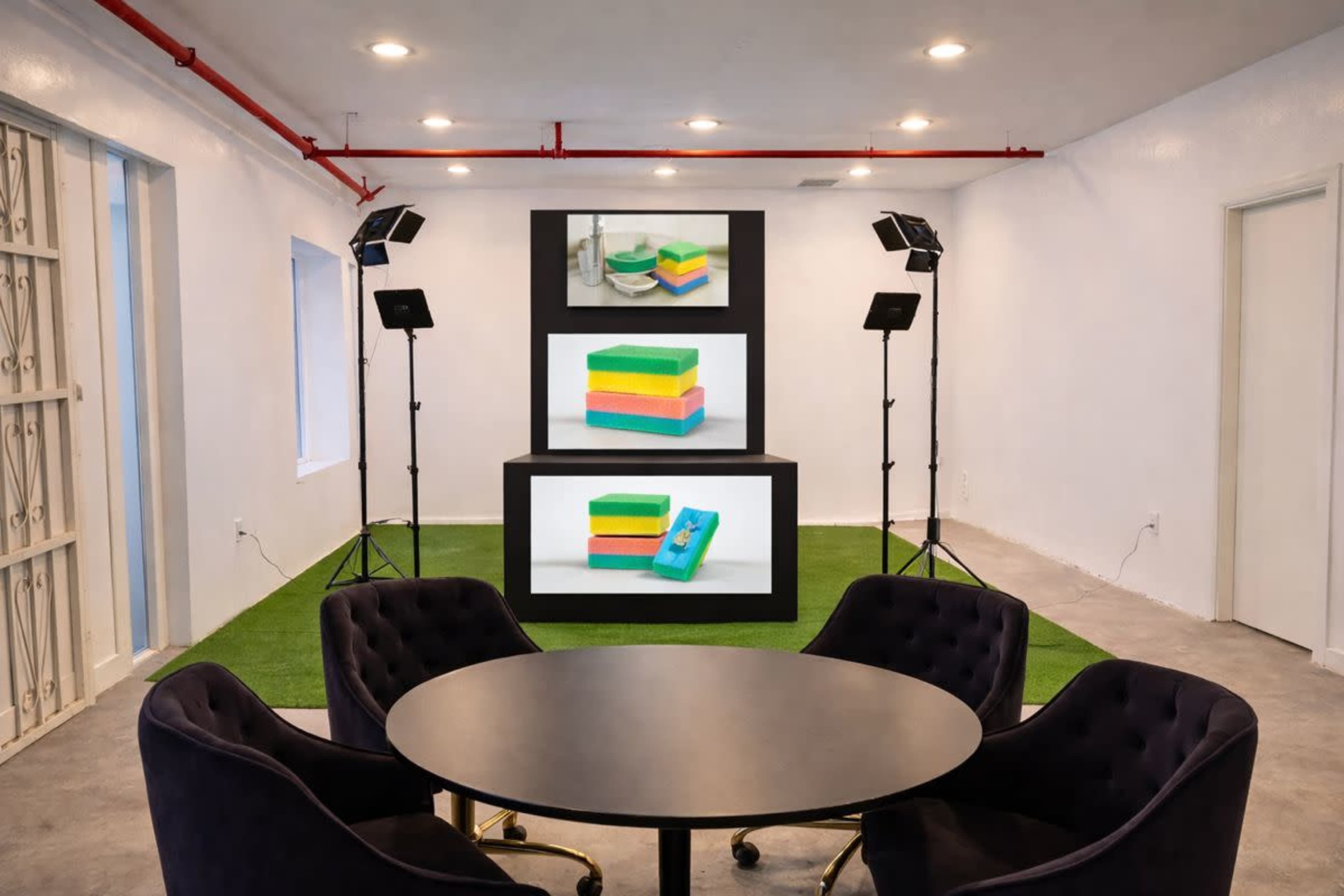 The image shows a bright, minimalist room with a round black table and plush chairs, featuring three stacked screens displaying colorful sponges.